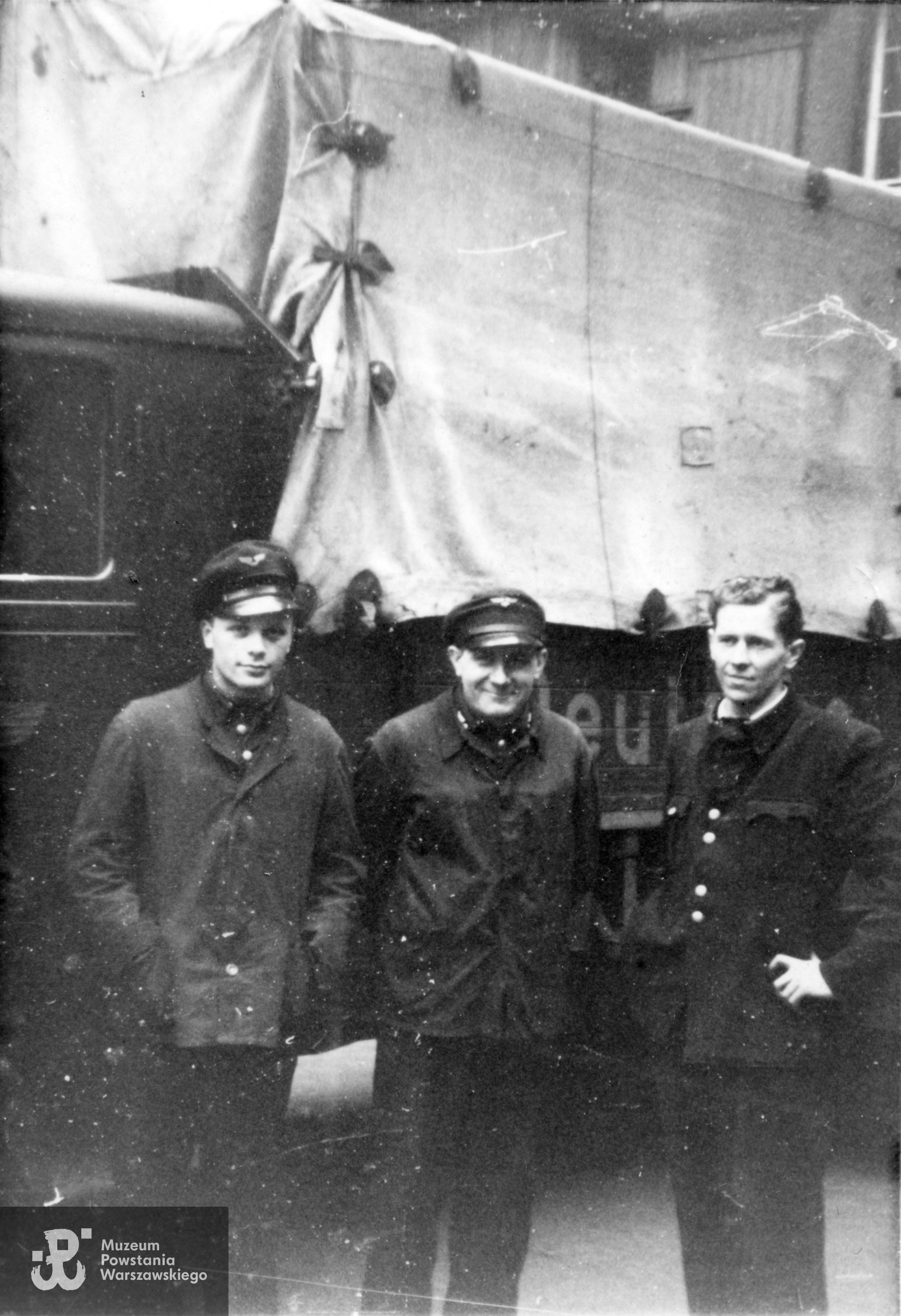 Fotografia z okresu okupacji niemieckiej. Trzej młodzi mężczyźni w  mundurach,  najprawdopodobniej pracownicy Deutsche Post stojący na tle częściowo widocznej ciężarówki z plandeką i częściowym napisem  (D)EUT(SCHE). Pierwszy z prawej  stoi Zygmunt Biernacki "Sęk". Fot. ze zbiorów Muzeum Powstania Warszawskiego, sygn. MPW-IP/9406 (P/3557).