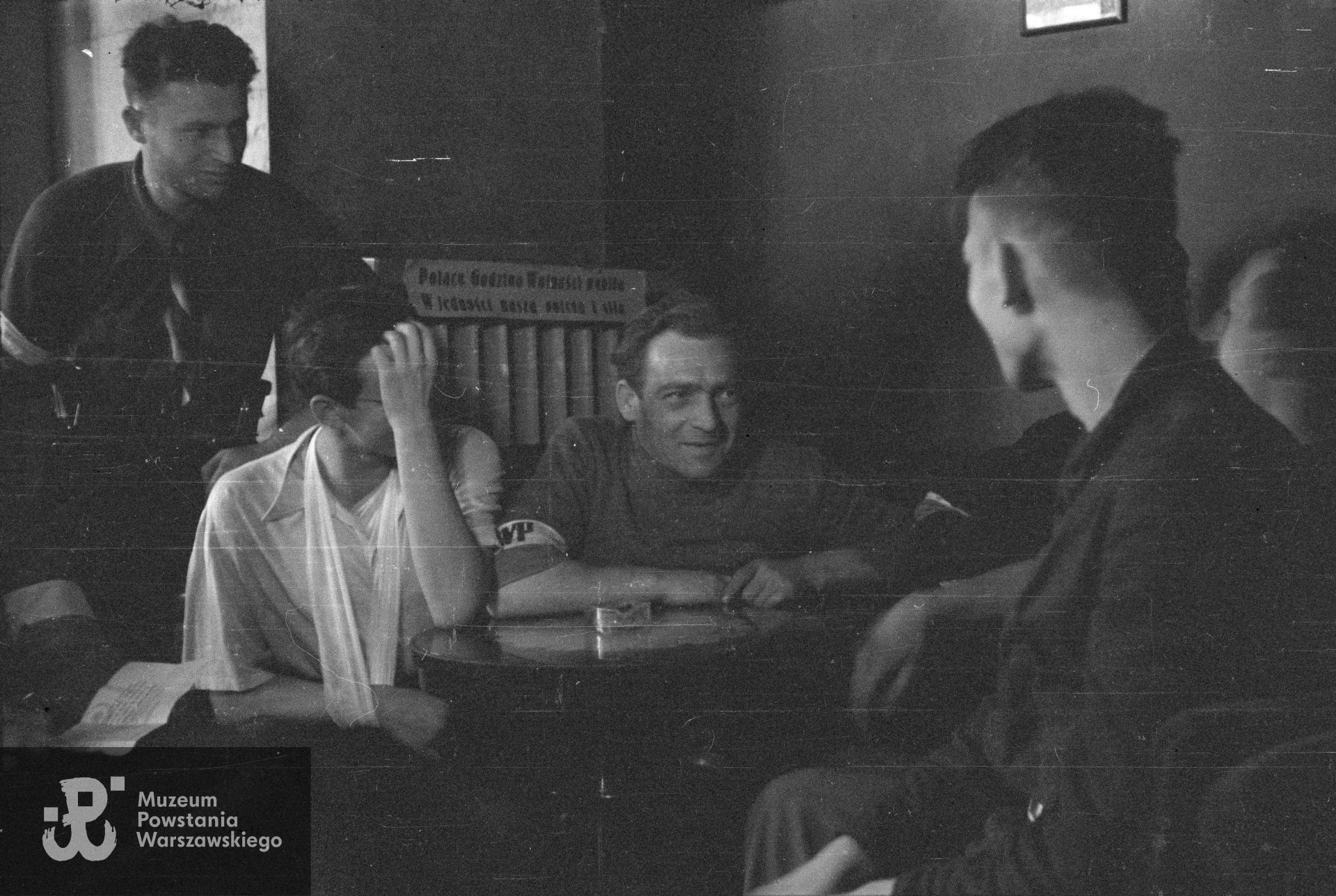Fotografia z Powstania Warszawskiego. Śródmieście Północne. Żołnierze z Oddziału Osłonowego Wojskowych Zakładów Wydawniczych (WZW) w powstańczej kantynie, utworzonej w kawiarni Yacht-Klub w kamienicy Lewenfisza przy ulicy Sienkiewicza 1. Od lewej stoi: plut. Wacław Latoszek "Kukułka"; siedzą: kpr. pchor. Wojciech Szendzikowski "Marabut" i por. Stefan Berent "Steb". Autorem zdjęcia jest Eugeniusz Lokajski „Brok”. Fot. ze zbiorów Muzeum Powstania Warszawskiego, sygn. MPW-IN/233