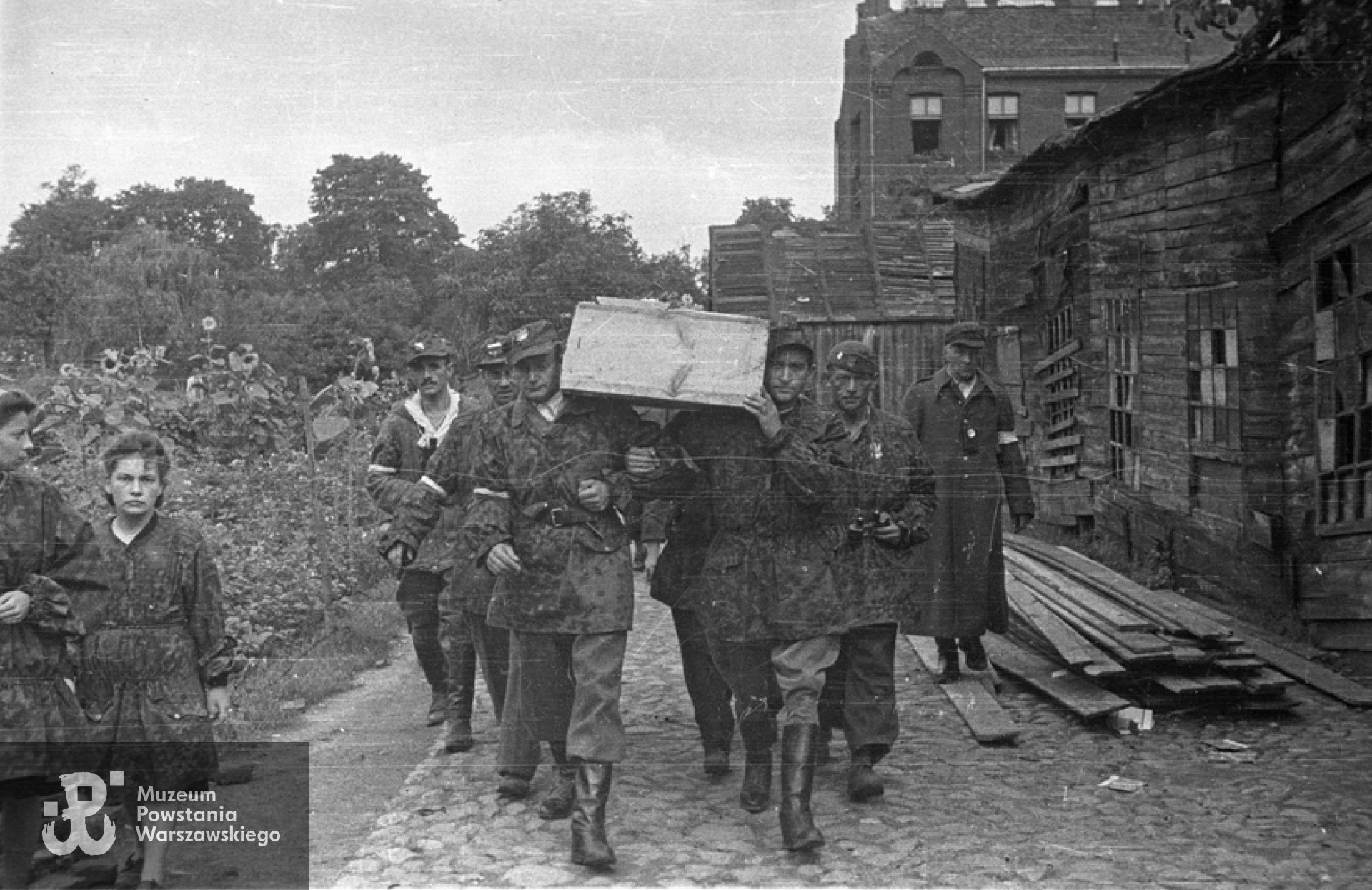 Fotografia z Powstania Warszawskiego. Wola. Żołnierze kompanii "Zemsta" batalionu "Pięść" niosący trumnę poległego ppor. Tadeusza Towarnickiego "Naprawa". Trumnę niosą ppor. Kazimierz Barszczewicz "Henryk" i ppor. "Ryś"(Jan Nowicki). Z lewej widoczny ppor. Jan Kowalczyk "Kieł", z lornetką po prawej Eugeniusz Kuźwa "Hipek" dowódca 169 plutonu batalionu "Kiliński". W tle po prawej widoczny budynek Zakładu dla umysłowo niedorozwiniętych na posesji Karolkowa 77. Autorem zdjęcia jest Stefan Bałuk "Starba", "Kubuś" Fot. ze zbiorów Muzeum Powstania Warszawskiego, sygn. MPW-IS/149