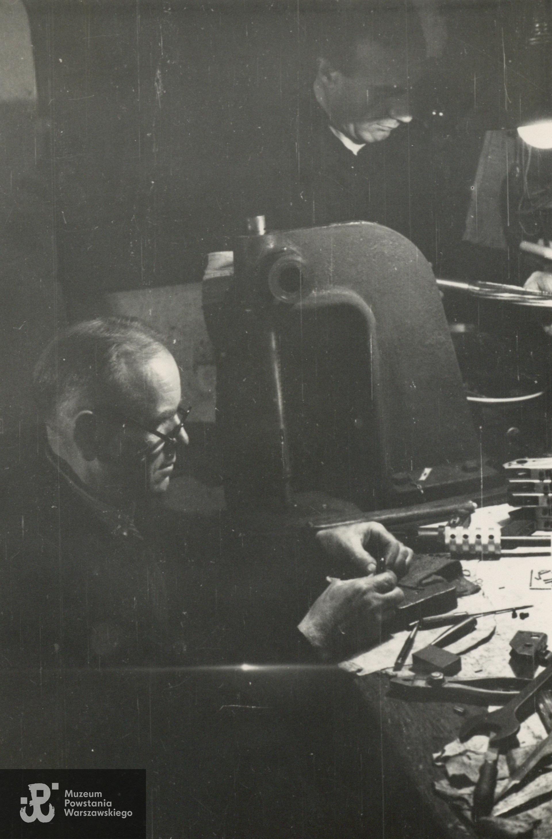 Fotografia z Powstania Warszawskiego. Śródmieście Północne. Montownia pistoletów maszynowych Błyskawica w kamienicy Lewenfisza przy pl. Napoleona 1, róg Boduena 2, róg Sienkiewicza 1. Rusznikarze z Oddziału Osłonowego Wojskowych Zakładów Wydawniczych (WZW) podczas pracy. Przy stole siedzi sierż. Roman Brzosko "Szpak" , w głębi stoi st. wachm. Roch Kafarowski "Ksawery"   Fot. ze zbiorów Muzeum Powstania Warszawskiego, sygn. P/7958, ujęcie tożsame: MPW-IN/367. Autorem zdjęcia jest Eugeniusz Lokajski "Brok"
