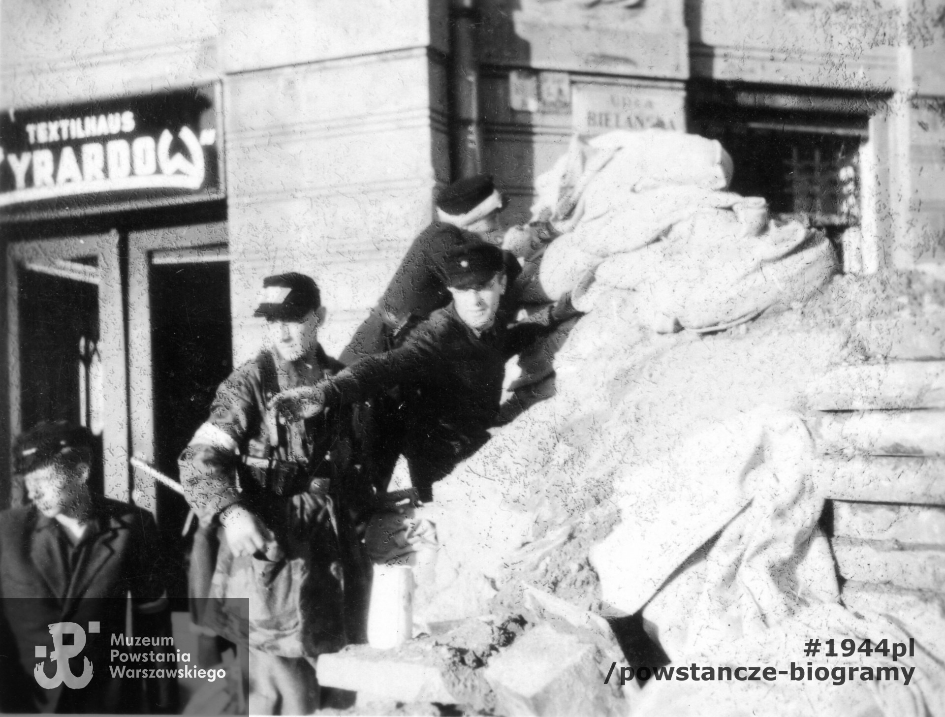 Fotografia z Powstania Warszawskiego. Starówka. Powstańcy przy barykadzie „Żyrardów”, zwanej też redutą „św. Trójcy” u wylotu ul. Daniłowiczowskiej na Bielańską. W tle widoczne wejście do sklepu firmowego fabryki tekstyliów „Żyrardów” przy ul. Bielańskiej 8.  Autorem zdjęcia jest Edward Tomiak. Fot. ze zbiorów Muzeum Powstania Warszawskiego, sygn. MPW-IP/4945/101. Na zdjęciu widoczni - na dole po lewej plut. pchor. Mieczysław Sztejerwald ps. Mieczysław, nad nim dowódca barykady por. Jerzy Roman Dereń ps. „Zawrot" (1915-1986). Identyfikacja - p. Jacek Sztejerwald - <i>Powstanie Warszawskie - Batalion KB "Nałęcz"</i>
