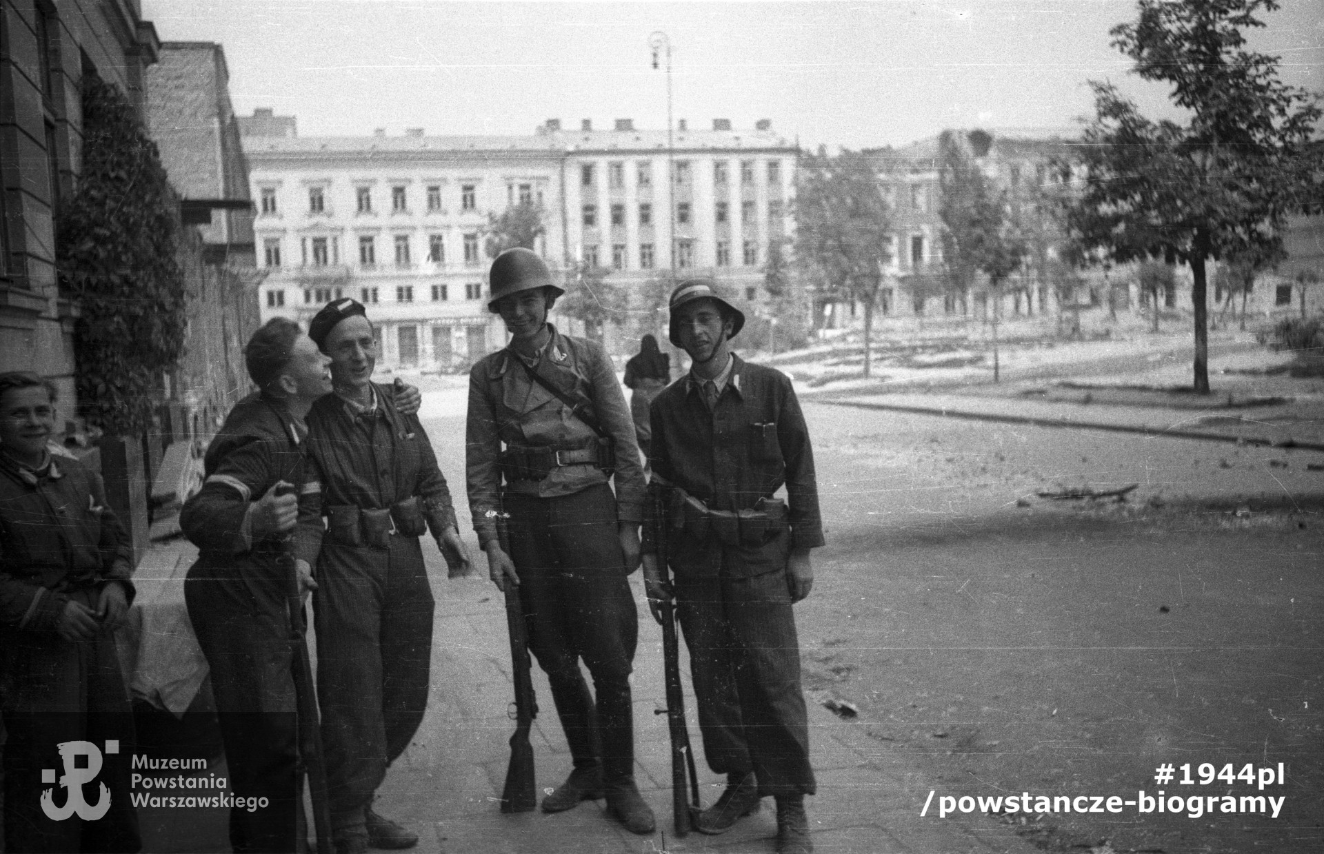 Fotografia z Powstania Warszawskiego. Śródmieście Północne. Pięciu żołnierzy 1150 plut. Zgrupowania „Bartkiewicz” (trzej uzbrojeni z karabiny) przed swoją kwaterą na pl. Dąbrowskiego 5. Ujęcie w kierunku północnym. W tle przechodząca kobieta, prawdopodobnie siostra zakonna.  Autorem zdjęcia jest Joachim Joachimczyk. Fot. ze zbiorów Muzeum Powstania Warszawskiego,  sygn.  MPW-IN/9725. Kpr. pchor.  Józef Rumiński „Spartanin" stoi w środku.