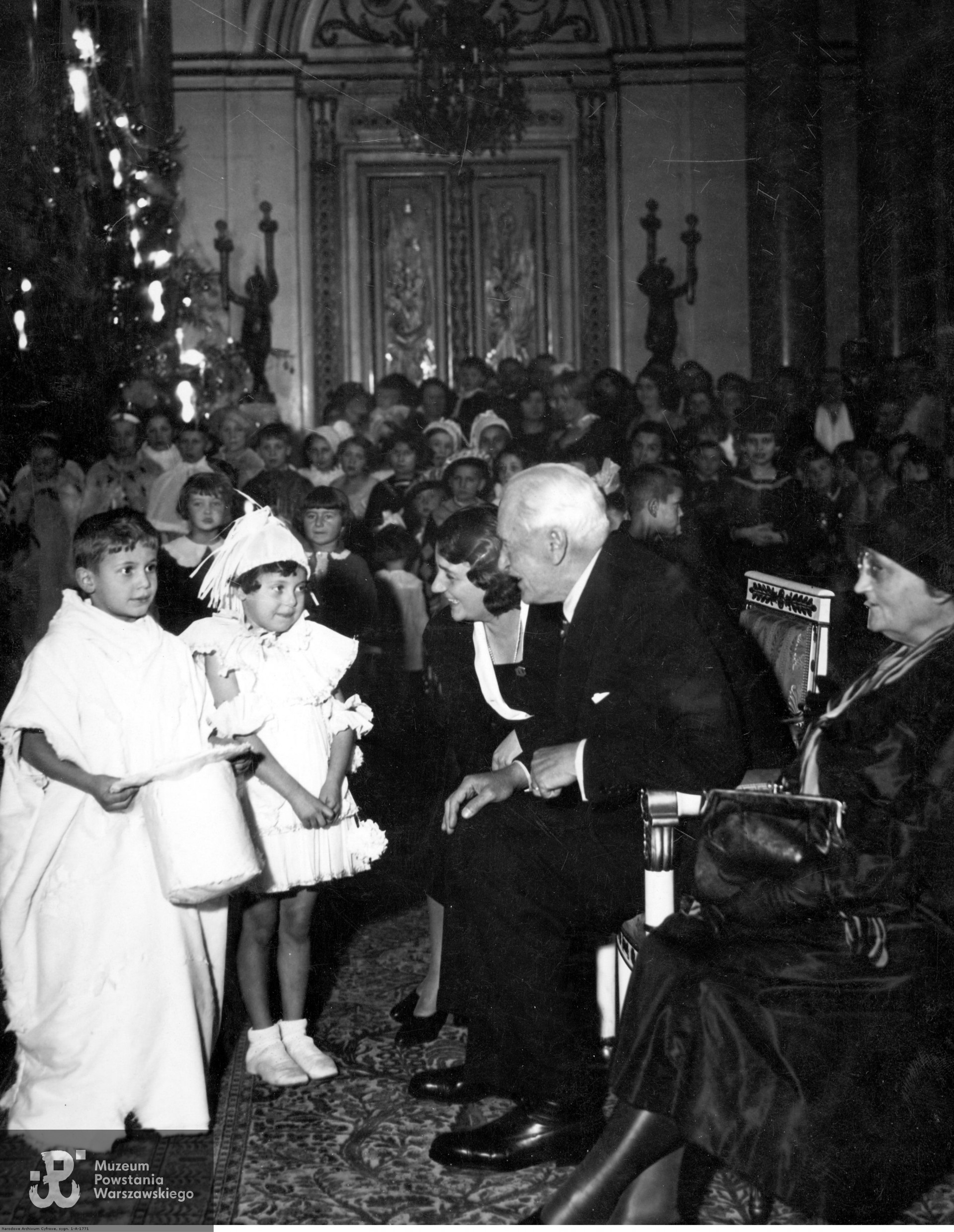  Grudzień 1934 roku.  Zabawa choinkowa dla dzieci funkcjonariuszy zamkowych na Zamku Królewskim w Warszawie. Prezydent RP Ignacy Mościcki z żoną Marią wśród dzieci - na pierwszym planie od lewej - Jerzy Albin Olk, Niusia Oleszczuk. Źródło: zbiory NAC, Koncern Ilustrowany Kurier Codzienny - Archiwum Ilustracji, Sygnatura: 1-A-1771.