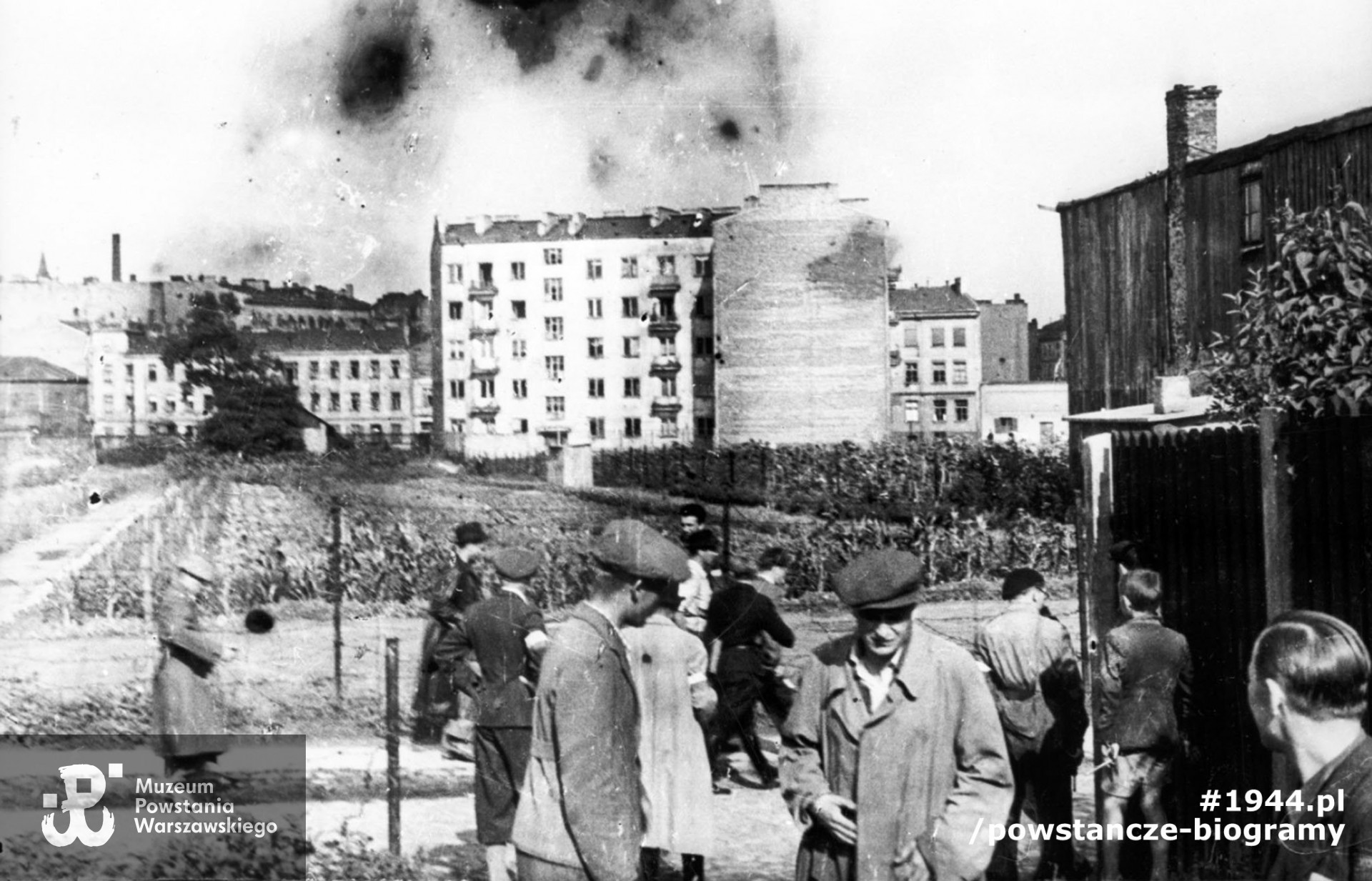 Wola. Godzina "W". Grupa powstańców z Batalionu "Pięść" stojąca przy wylocie ul. Długosza na Karolkową, oczekująca na koncentrację w rejonie cmentarza ewangelicko-augsburskiego. Na drugim planie, tyłem, ubrany w spodnie pumpy, stoi mjr Alfons Kotowski "Okoń", dowódca Batalionu, obok w jasnym płaszczu ppor. Stanisław Bąk "Burza". Ujęcie w kierunku wschodnim. W tle widoczne budynki przy zbiegu ul. Okopowej i ul. Wolność; po lewej kamienica przy Okopowej 30 róg Wolność 21, w środku czteropiętrowa kamienica przy Okopowej 33. Autorem zdjęcia jest Stefan Bałuk. Ze zbiorów Muzeum Powstania Warszawskiego, sygn. MPW-IH/3412. Prawa autorskie MPW