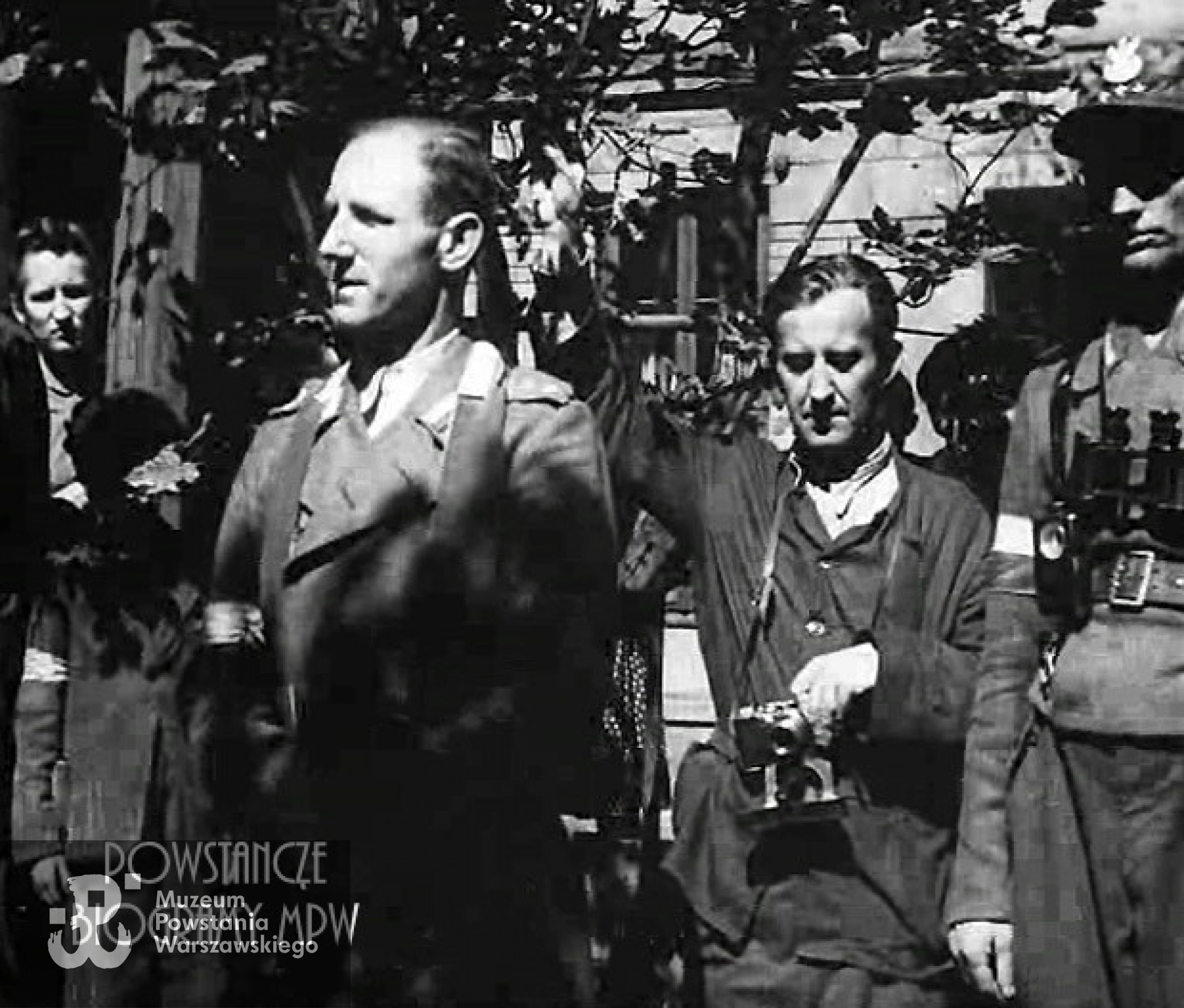 Zdjęcie filmowe w powstańczej kroniki. Na pierwszym planie przemawiający ksiądz mjr. Władysław Zbłowski ps. "Struś" Na drugim planie z aparatem fotograficznym na szyi szef operatorów w Wydziale Propagandy Oddziału II Sztabu KG AK reżyser Antoni Bohdziewicz „Wiktor”. Panorama w prawo na kpt. „Hala” i rodzinę poległego łącznika. Ze zbiorów Muzeum Powstania Warszawskiego: <i>powstaniewarszawskiefilm.pl</i>