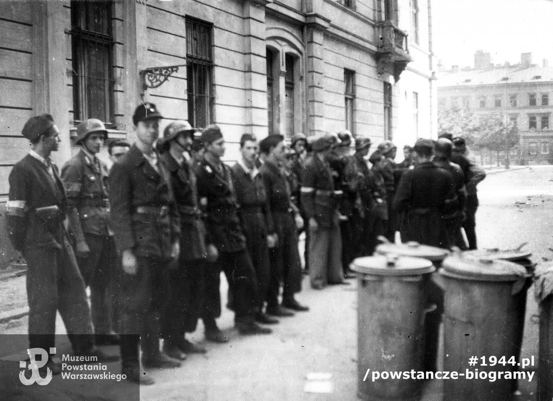 Fotografia z Powstania Warszawskiego. Śródmieście Północne. Zbiórka plutonu "Rygla" zgrupowania "Bartkiewicz" przy ul Rysiej przed akcją w Ogrodzie Saskim. Ujęcie w kierunku wschodnim. Za plecami powstańców elewacja dawnego Hotelu Francuskiego - Marszałkowska 146 róg Rysia róg pl. Dąbrowskiego 5. W tle po prawej kamienica przy pl. Dąbrowskiego 2/4. Pierwszy rząd: Janusz Sawicki "Zabawa", "Władek", "Chimera", "Krakus". Drugi rząd: "Korczak", "Bohdan", "Leszczyc", NN, "Kalinowski", "Sęp"Ze zbiorów Muzeum Powstania Warszawskiego, sygn. MPW-IP/3843