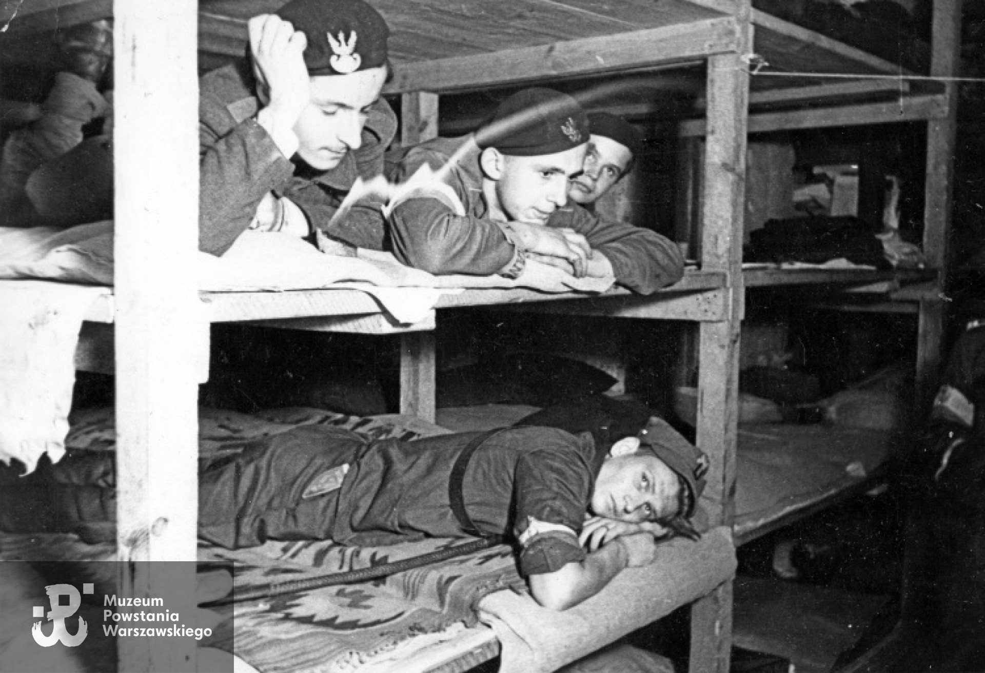 27 sierpnia 1944. Sypialnia plutonu 569 (z 2 Harcerskiej Baterii Artylerii Przeciwlotniczej "Żbik" Zgrupowanie "Gurt") na ul. Chmielnej 61. Śródmieście Północne. Na górze od lewej: 1. kanonier Janusz Preyss "Kowalski" 2. Kazimierz Rakowski "Świteź" 3. Andrzej Mieszczański "Hetman" Na dole kanonier Ryszard Głuchowski "Jaworzak". Półki na których składano papier służą za łóżka. Autorem zdjęcia jest Joachim Joachimczyk. Zbiory MPW, sygn. MPW-IK/1813. Prawa autorskie: MPW