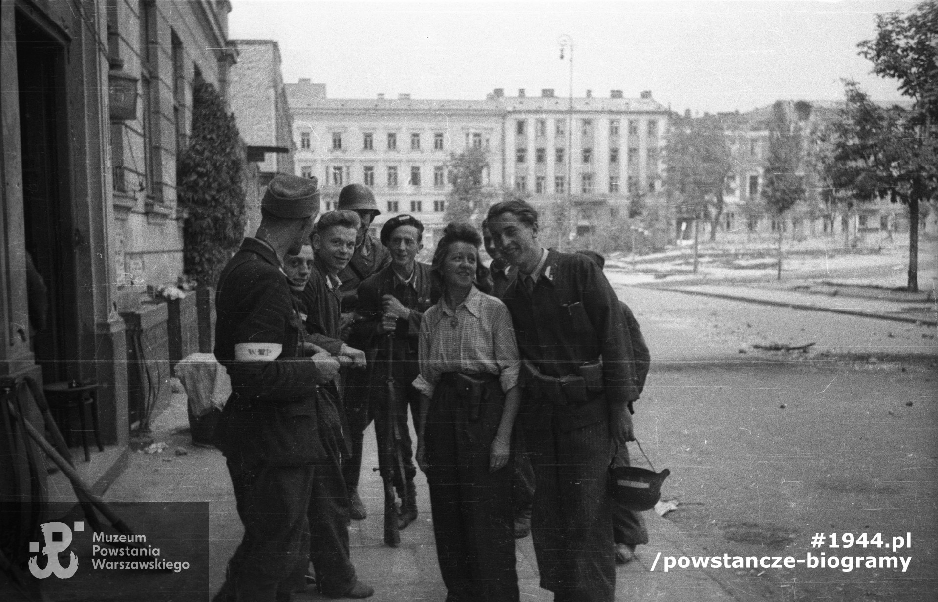 Fotografia z Powstania Warszawskiego. Śródmieście Północne. Dowódca plutonu 1150 por. Władysław Darmosz "Kutno" (pierwszy od lewej) wśród swoich żołnierzy przed kwaterą na pl. Dąbrowskiego 5. Ujęcie w kierunku północnym. Autorem zdjęcia jest Joachim Joachimczyk "Joachim". Fot. ze zbiorów Muzeum Powstania Warszawskiego, sygn. MPW-IN/9727