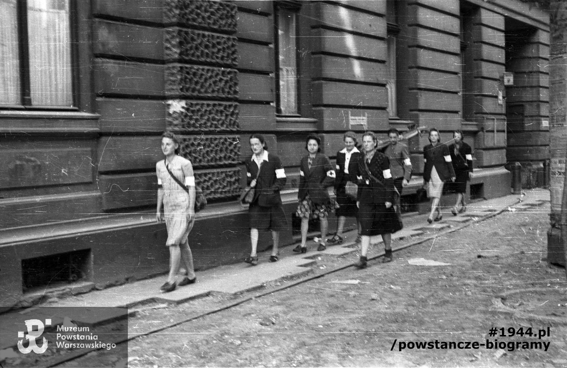 Fotografia z Powstania Warszawskiego. Śródmieście Północne. Grupa sanitariuszek z kompanii „Koszta” przechodząca obok domu przy Moniuszki 9; od lewej: Gabriela Karska „Joasia”, dr Zofia Lejmbach „Róża”, Danuta Laskowska „Dorota”, (NN) „Małgorzata”, Halina Wojtczuk „Halszka”, Wanda Regenciak „Alicja”, dr Danuta Staszewska „Marta”, z boku idzie Melania Wolska „Ewa Samarytanka”. Autorem zdjęcia jest Eugeniusz Lokajski „Brok". Fot. ze zbiorów Muzeum Powstania Warszawskiego, sygn. MPW-IN/47
