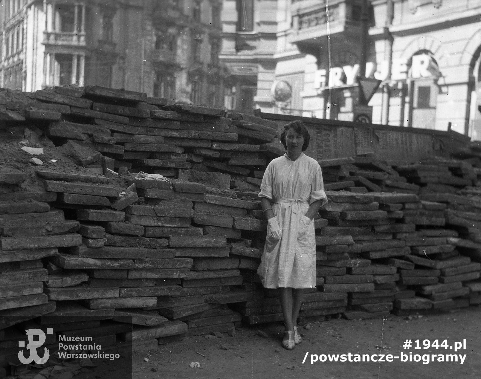 Fotografia z Powstania Warszawskiego. Zdjęcie wykonane na skrzyżowaniu ul. Koszykowej z ul. Marszałkowską. Sanitariuszka szpitala przy ul. Marszałkowskiej 40 Wanda Srzednicka "Aza" przy barykadzie z płyt chodnikowych przegradzającej ul. Koszykową wzdłuż ul. Marszałkowskiej (pomiędzy kamienicami Marszałkowska 46 i Marszałkowska 48). Nad barykadą widoczna kamienica Marszałkowska 48 (róg Koszykowa). Po prawej widoczny fragment kamienicy przy Marszałkowskiej 48 (widoczna elewacja od strony Koszykowej z neonem "Fryzjer"). Autorem zdjęcia jest Zofia Wolska-Degórska. Fot. ze zbiorów Muzeum Powstania Warszawskiego, sygn. MPW-IN/10918 