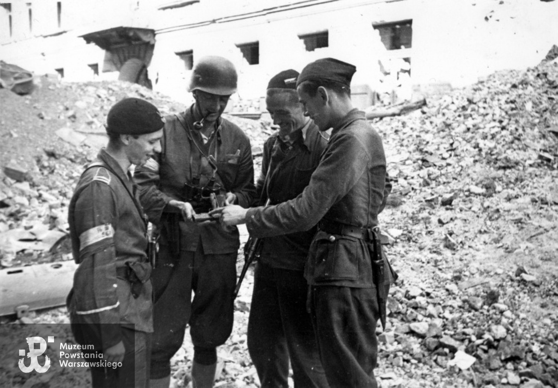 27 sierpnia 1944 roku, powstańcy oglądający pistolet. Od lewej: kapral podchorąży Adam Bóbr ps. "Jaxa", plutonowy Włodzimierz Dąbkowski ps. "Michał Romer", sierżant Zygmunt Klimowicz ps. "Neron" i podchorąży Marek Raczyński ps. "Żwir". Zdjęcie wykonane przed kwaterą przy ul. Chmielnej 61. Po lewej stronie widoczna barykada od strony ul. Żelaznej. Fot. Joachim Joachimczyk. Zbiory fototeki MPW, sygnatura MPW-IP/2678, prawa autorskie MPW. Zdjęcie dostępne także jako Archiwum Fotograficzne Stefana Bałuka, zbiory NAC, sygn. 37-1539-2