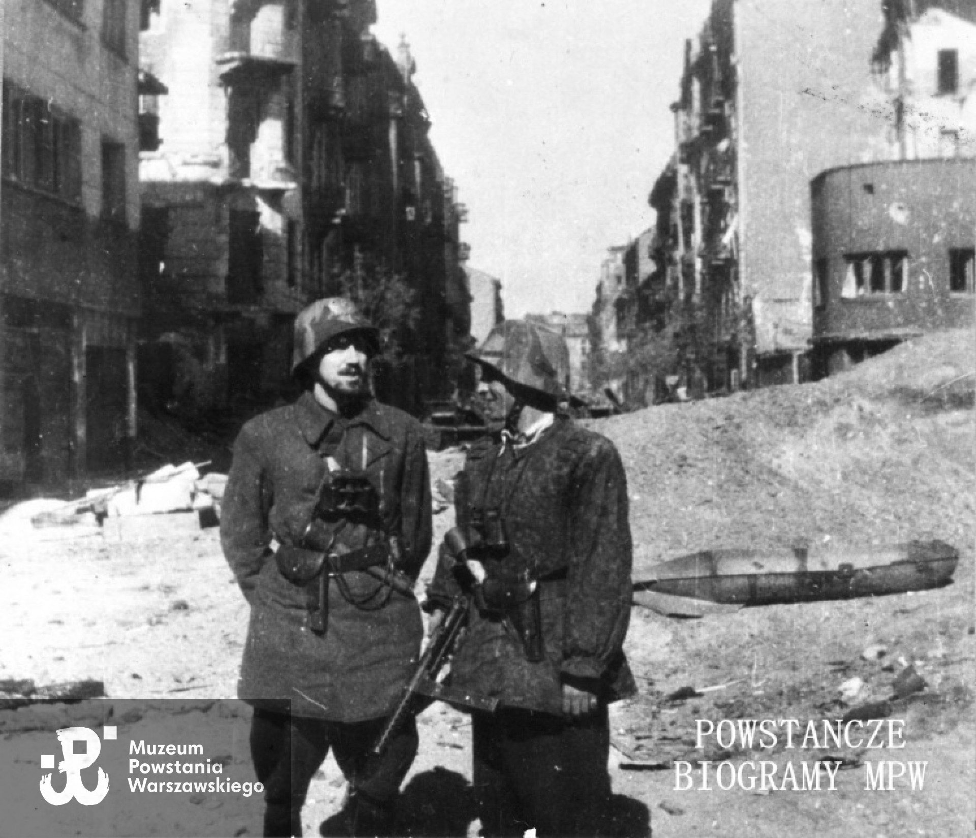 Fotografia z Powstania Warszawskiego. Śródmieście Północne. Dwóch powstańców ze zgrupowania "Chrobry II" na ul. Miedzianej. Po lewej por. Zbigniew Brym "Zdunin" z I batalionu "Lecha Żelaznego" 3 kompania. Ujęcie od strony ul. Srebrnej w kierunku północnym. Na drugim planie po prawej obudowa niemieckiej bomby kastowej AB 250. W tle od lewej: budynek Srebrna 1, wylot ul. Srebrnej, narożna kamienica Miedziana 1 / Srebrna 6 a dalej domy Srebrna 3, 5 i 7, Od prawej: piętrowy budynek Miedziana 2 / Srebrna 4, ruiny kamienicy Miedziana 4, a dalej domy Miedziana 4a, 6, 8 i kolejne. Fot. ze zbiorów Muzeum Powstania Warszawskiego, sygn. MPW-IK/1909