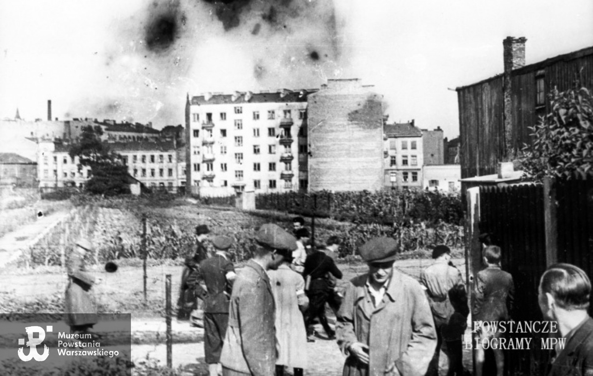 Ta sama fotografia ze zbiorów Muzeum Powstania Warszawskiego, opis:  Wola. Godzina "W". Grupa powstańców z Batalionu "Pięść" stojąca przy wylocie ul. Długosza na Karolkową, oczekująca na koncentrację w rejonie cmentarza ewangelicko-augsburskiego. Na drugim planie, tyłem, ubrany w spodnie pumpy, stoi mjr Alfons Kotowski "Okoń", dowódca Batalionu, obok w jasnym płaszczu ppor. Stanisław Bąk "Burza". Ujęcie w kierunku wschodnim. W tle widoczne budynki przy zbiegu ul. Okopowej i ul. Wolność; po lewej kamienica przy Okopowej 30 róg Wolność 21, w środku czteropiętrowa kamienica przy Okopowej 33.Autorem zdjęcia jest Stefan Bałuk. Ze zbiorów Muzeum Powstania Warszawskiego, sygn. MPW-IH/3412. Prawa autorskie MPW