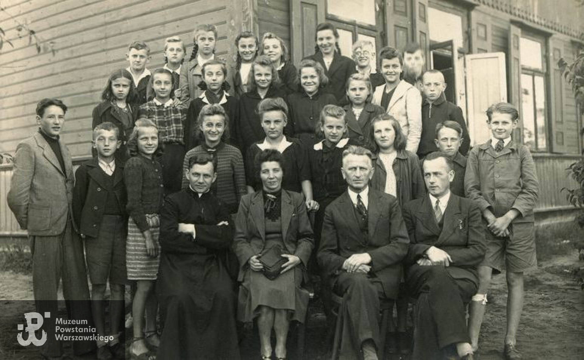 Fotografia z okresu okupacji niemieckiej przedstawiająca nauczycieli i uczniów Szkoły Powszechnej nr 1 w Legionowie. Ks. Wacław Szelenbaum siedzi w pierwszym rzędzie, pierwszy z lewej strony.  Fot. ze zbiorów Muzeum Historycznego w Legionowie, źródło: Gazeta Powiatowa, <i>Wojenne adresy Legionowa: Okupacyjna „Jedynka”</i>