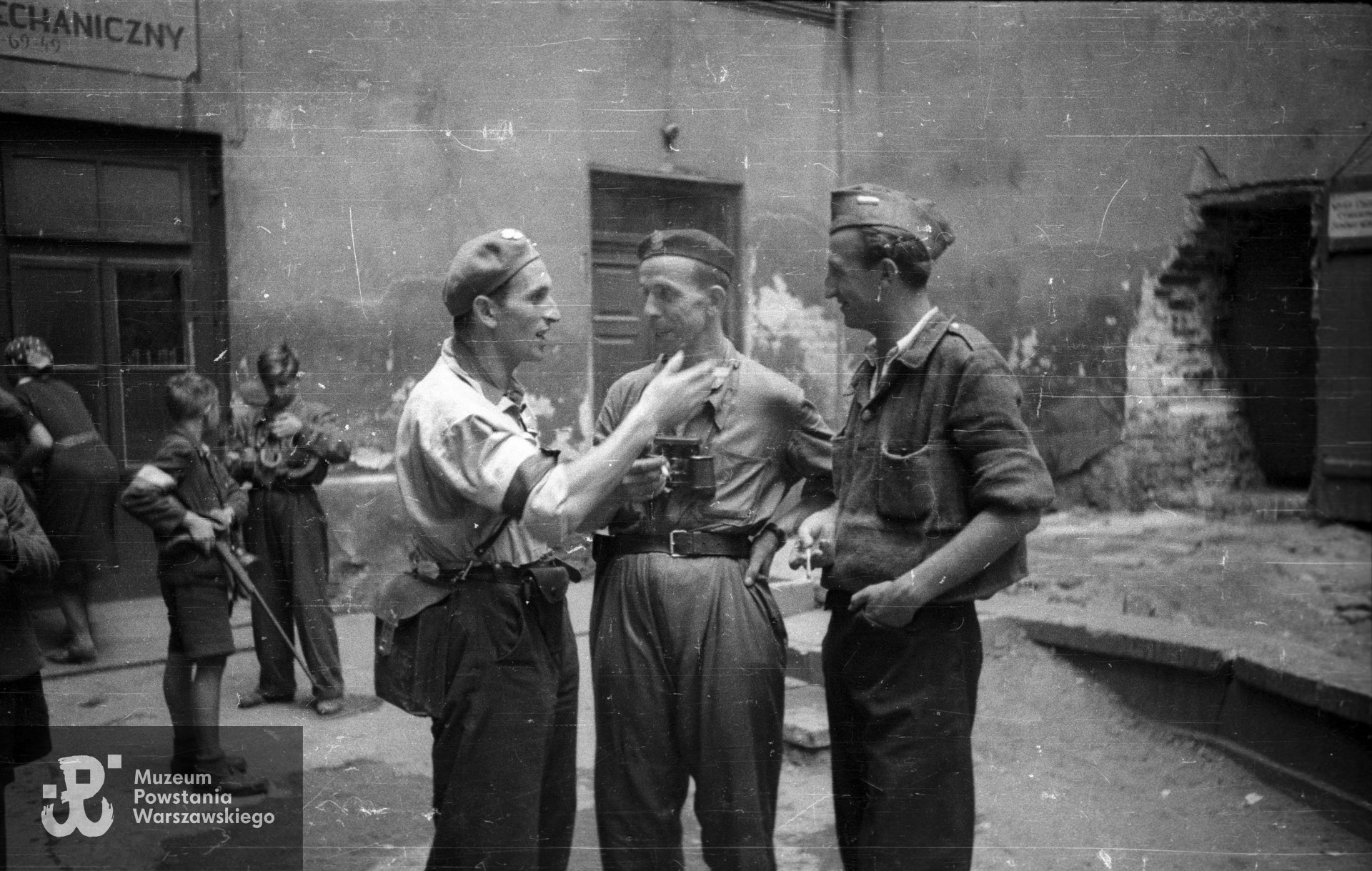 Fotografia z Powstania Warszawskiego. Śródmieście Południowe, prawdopodobnie drugie podwórko kamienicy przy Żurawiej 6. Fotoreporter Joachim Joachimczyk "Joachim" (pierwszy z lewej) w rozmowie z oficerami batalionu "Sokół". Z prawej ppor. Brunon Wolski "Goryl", w środku kpt. Alfons Laska "Skała". Fot. ze zbiorów Muzeum Powstania Warszawskiego, sygn.  MPW-IN/10517, ujęcie tożsame MPW-IV/1876