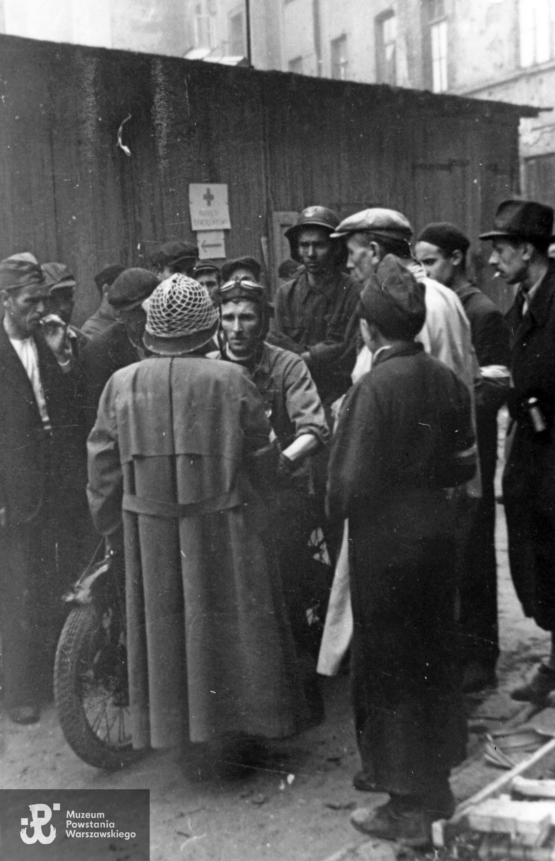 Fotografia z Powstania Warszawskiego. Śródmieście Północne. Grupa powstańców ze Zgrupowania "Chrobry II" na podwórzu kamienic przy ul. Twardej 40 i 38. Na motocyklu siedzi plut. pchor. Kazimierz Szczeszek "Adam" z kompanii łączności.  Odwrócony tyłem stoi szef sztabu i zastępca dowódcy Zgrupowania "Chrobry II"  kpt. Jan Jaroszek ps. Proboszcz. Fot. ze zbiorów Muzeum Powstania Warszawskiego, sygn. MPW-IH/910. Autorem zdjęcia jest Jan Żułma ps. Felix