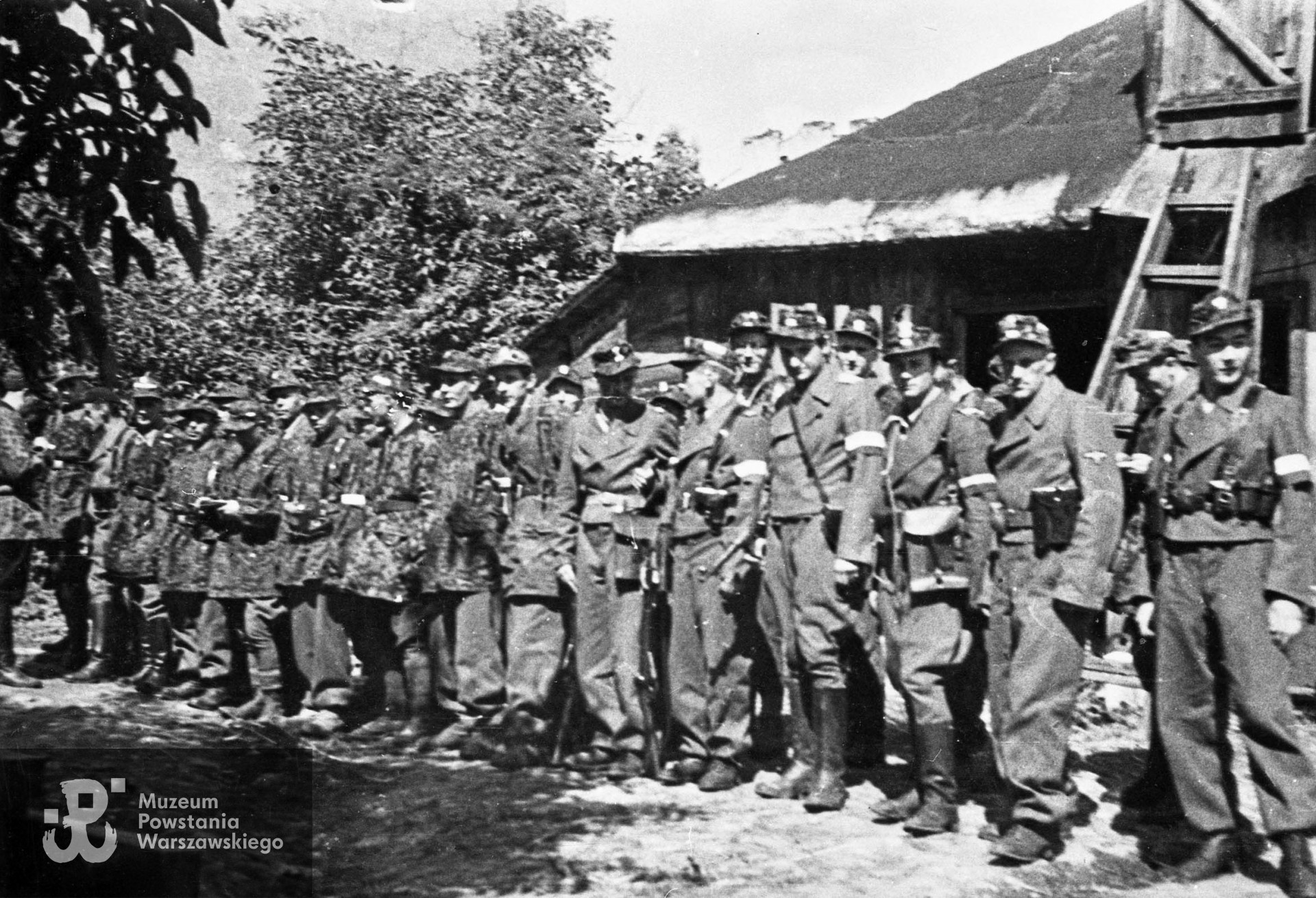 Fotografia z Powstania Warszawskiego. Wola. Żołnierze Kolegium "A" Kedywu OW AK w ogrodzie RGO - ul. Okopowa 41. Stoją od lewej: Aleksander Kabański "Brzozowski" (bokiem), trzeci - Jan Bagiński "Socha" (w berecie), czwarty - Zygmunt Siennicki "Bor" , ósmy - Józef Tomaszewski "Klemens". Dziewiąty od prawej - Jerzy Krzymowski "Jodła" (twarz w cieniu), czwarty od prawej- Jan Kulczycki "Argos" (w długich butach). Fot. ze zbiorów Muzeum Powstania Warszawskiego, sygn. MPW-IN/3643