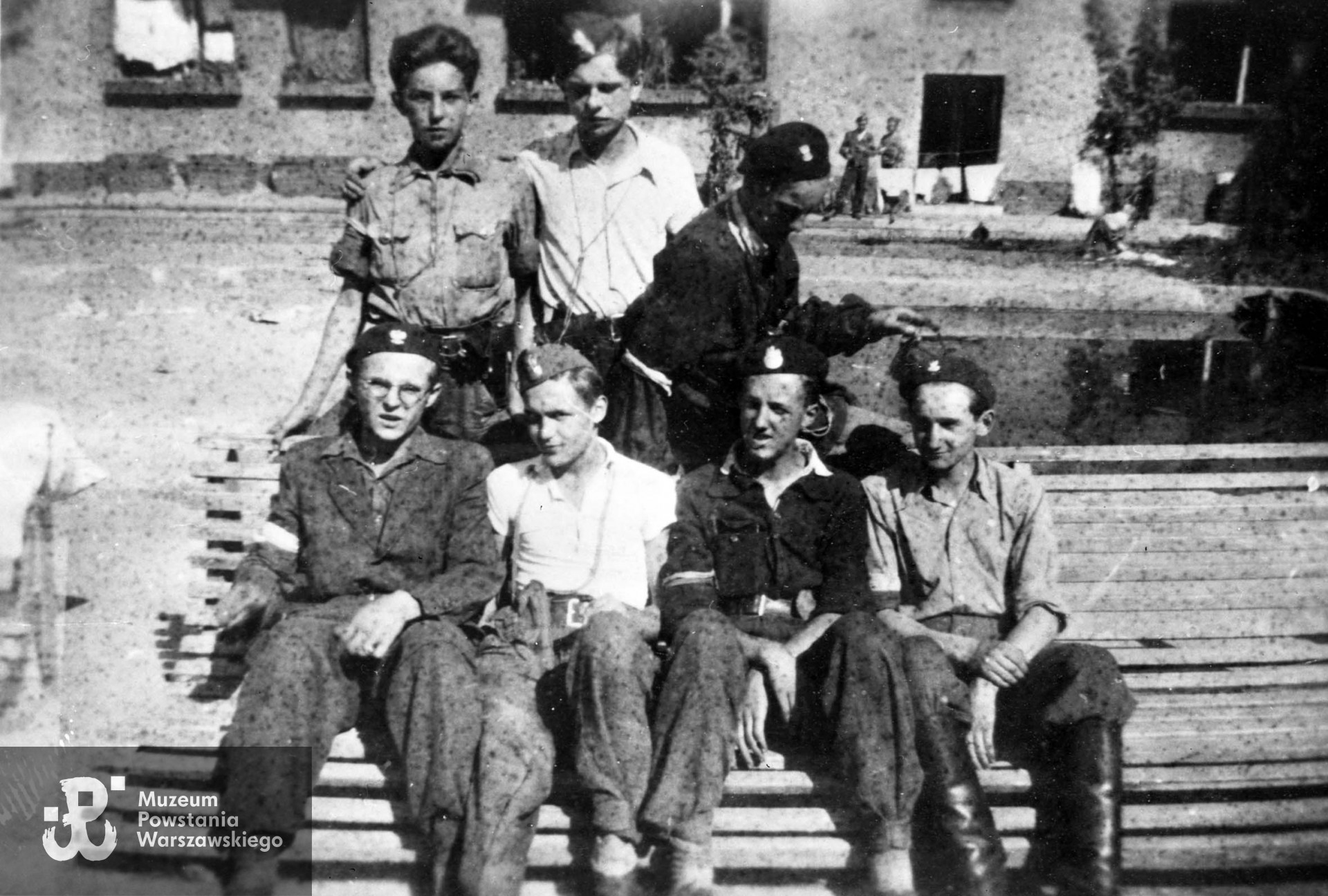 Fotografia z Powstania Warszawskiego. Żoliborz. Harcerze z plutonu 227, zgrupowania "Żyrafa" na podwórzu bloku przy ul. Krasińskiego 20. Od lewej stoją: strz. Włodzimierz Krystasiak "Mongoł", kpr. Janusz Makowski "Janusz", kpr. Waldemar Rolbiecki "Popielica". Od lewej siedzą: strz. Zbigniew Kamiński "Złamaniec", strz. Wiesław Nowak "Jastrząb", strz. Józef Miernowski "Ziuk", strz. Józef Małkowski "Sokół". Fot. ze zbiorów Muzeum Powstania Warszawskiego, sygn. MPW-IH/4891