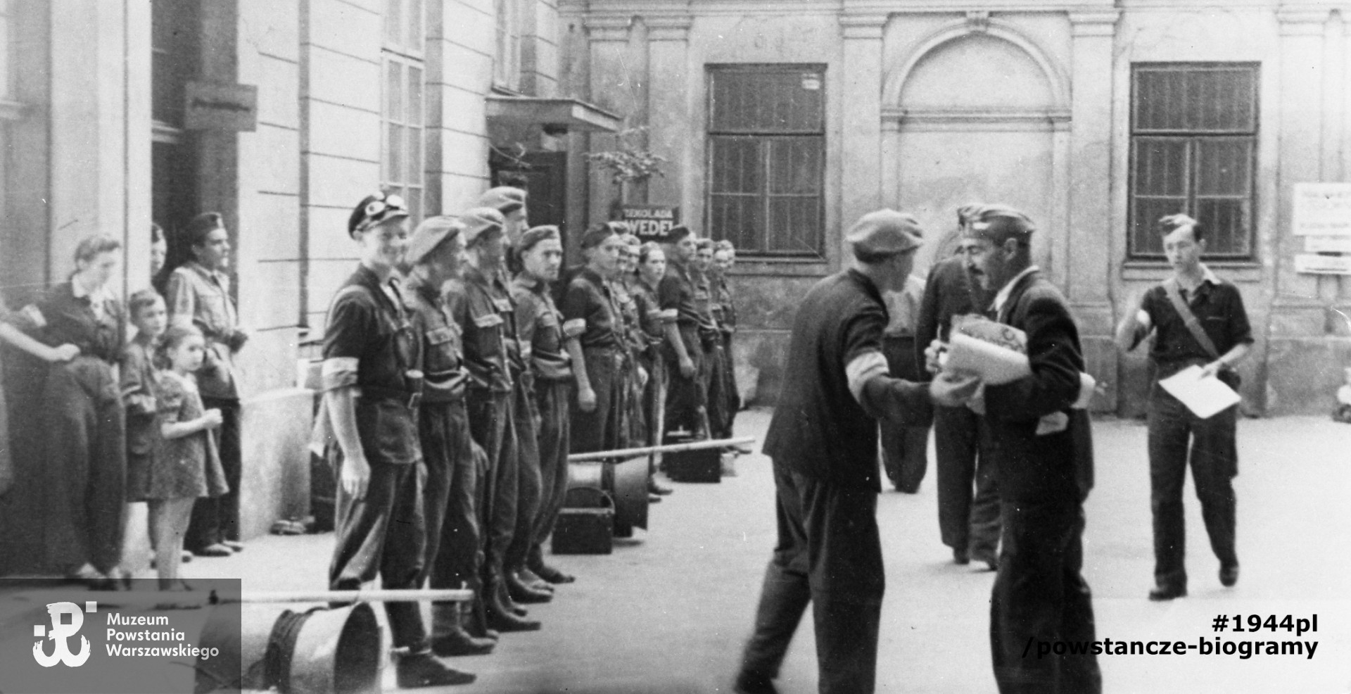 Fotografia z Powstania Warszawskiego. Śródmieście Północne. Odprawa oddziału tzw. "megafoniarzy" na podwórzu domu Wedla przy ul. Szpitalnej 8. Pierwszy od lewej Janusz Kulesza ps. "Super", dalej NN ps. "Wicher", Feliks Lendzion ps "Wołg". Po prawej trzymający pod pachą rulon papieru Ludwik Szumlewski ps "Selim". Fot. ze zbiorów Muzeum Powstania Warszawskiego,  sygn. MPW-IH/4878