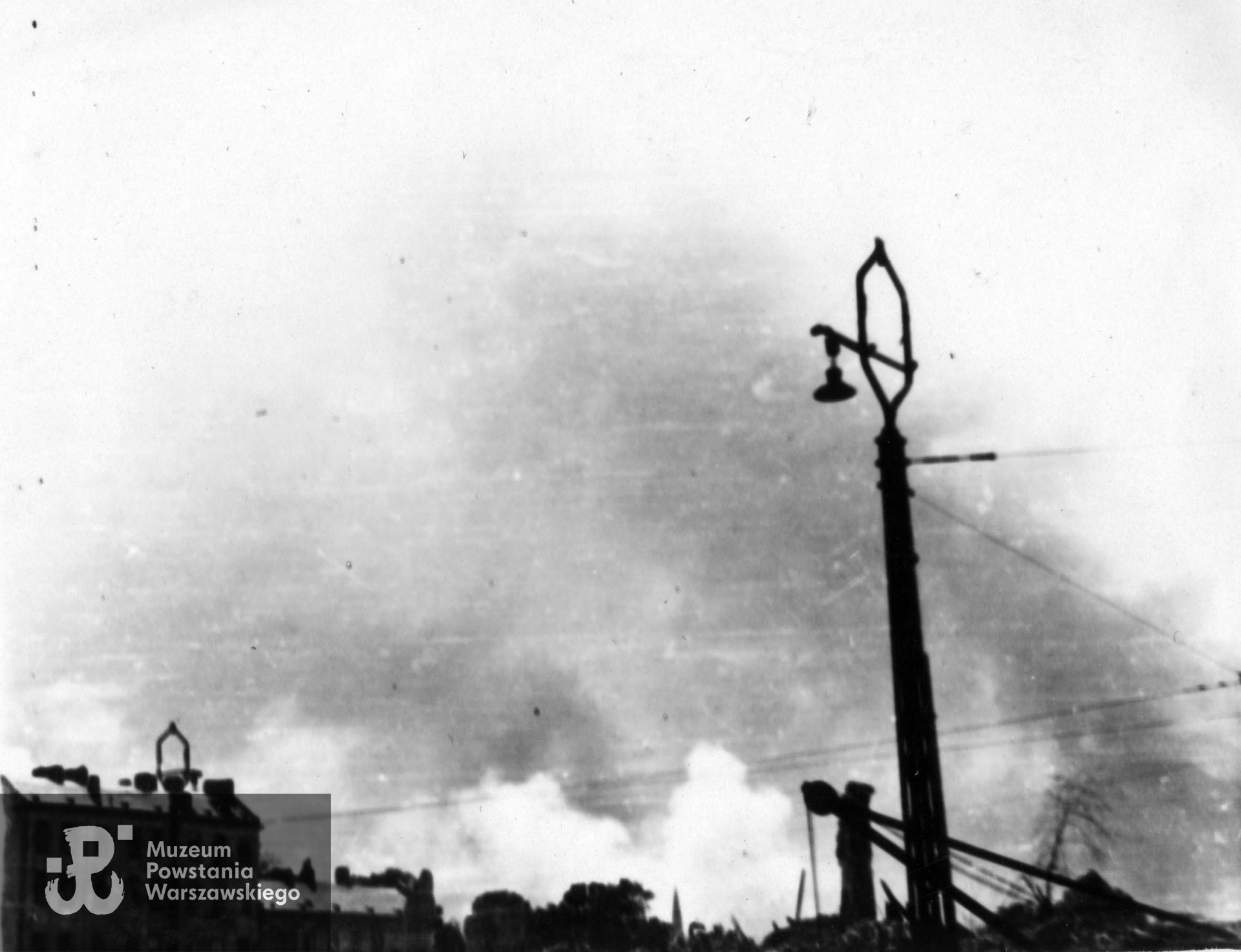 Fotografia autorstwa Zbigniewa Grochowskiego wykonana podczas Powstania Warszawskiego.  Dymy unoszące się nad miastem. Ujęcie z ulicy Bonifraterskiej w kierunku południowo-zachodnim. Po lewej widoczna kamienica przy ulicy Świętojerskiej 19, w środku między drzewami Ogrodu Krasińskich widoczna wieża kościoła ewangelicko-reformowanego na Lesznie. Fot. ze zbiorów Muzeum Powstania Warszawskiego, sygn. MPW-IP/2716 (MPW-GI-4000/15/06)