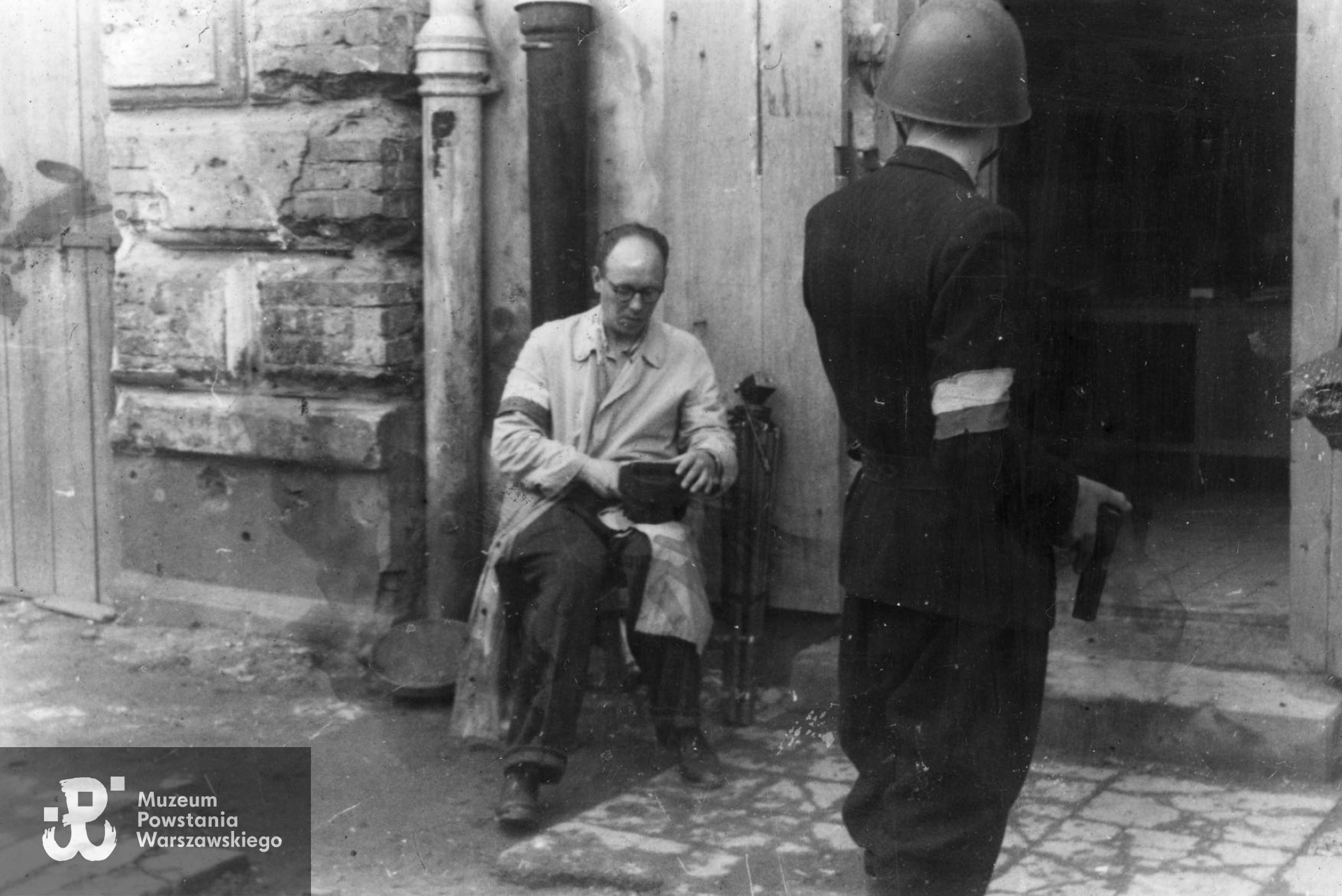 Fotografia z Powstania Warszawskiego autorstwa Bernarda Marwińskiego (PSW).  Śródmieście Północne, operator filmowy Henryk Vlassak siedzący na krześle przed wejściem do budynku. Na pierwszym planie tyłem Powstaniec z pistoletem. Fot. ze  zbiorów Muzeum Powstania Warszawskiego, sygn. MPW-IP/6831
