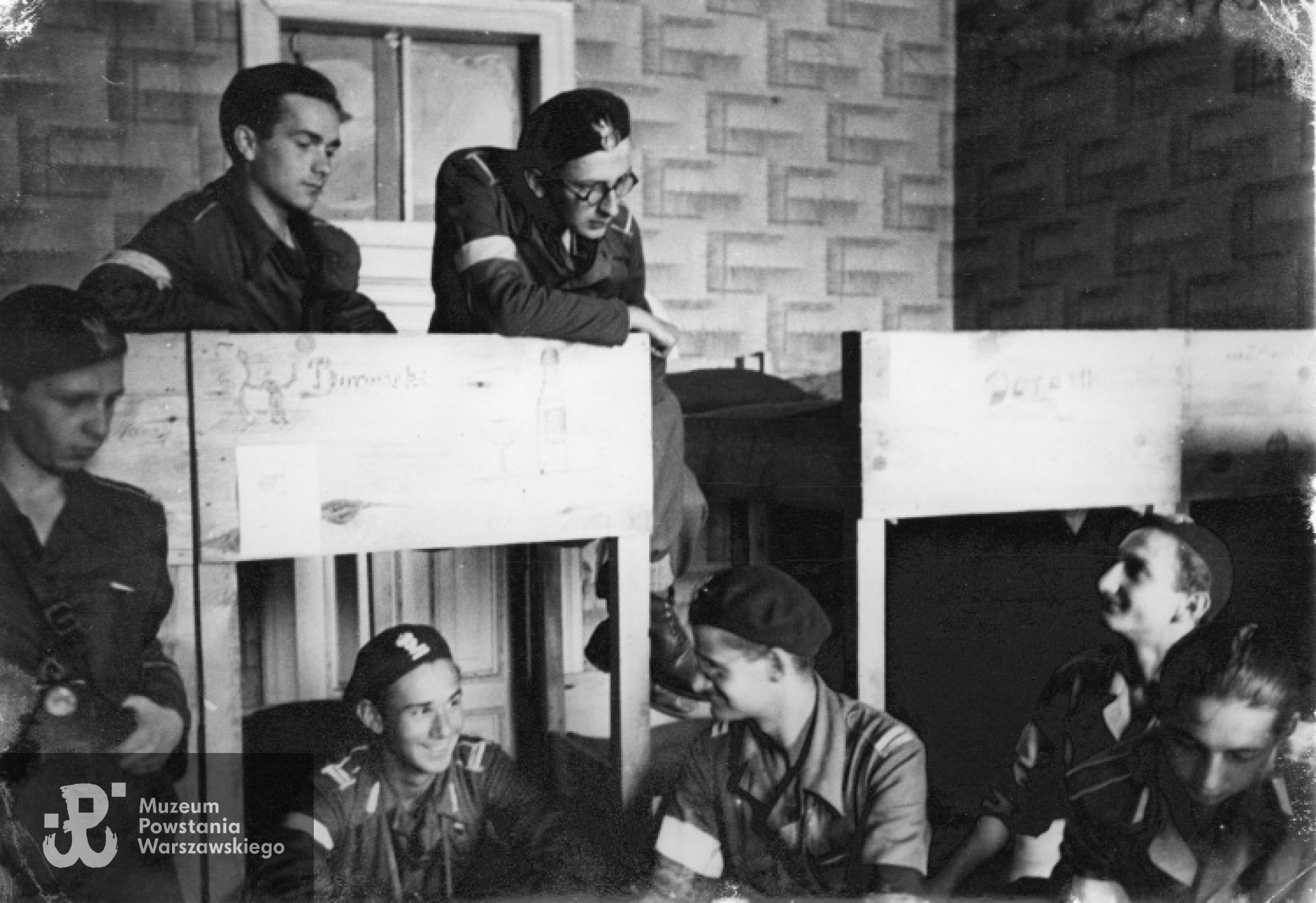 Fotografia z Powstania Warszawskiego - 27 sierpnia 1944 r. Powstańcy z plutonu 569 ppor. "Żuka" z 2 HBAP na kwaterze na Chmielnej 67. Od lewej stoi dowódca I plutonu Adam Bóbr "Jaxa", na dole siedzą: kpr. Marian Łączyński "Orlik", dowódca II plutonu Piotr Osiński "Kaczanowski", kan. Maciej Lewandowski "Michał", NN. Na górze na pryczach kan. Jan Urbański "Borowski" i bomb. Zbigniew Bartecki "Lubicz". Fotografia autorstwa Joachima Joachimczyka ps. "Joachim", zbiory fototeki MPW, sygn. MPW-IP/1251