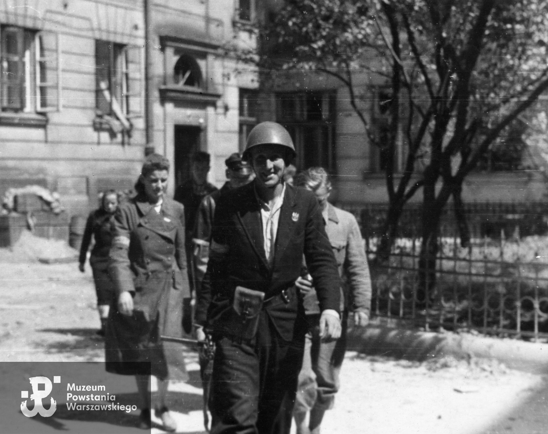 Fotografia z Powstania Warszawskiego. Grupa Powstańców z kompanii "Grażyna" Batalionu "Gustaw" idąca przez podwórze domu przy ulicy Mazowieckiej 4. Jako pierwszy idzie por. Władysław Wojewódzki "Kaszub". Ze zbiorów Muzeum Powstania Warszawskiego, sygn. MPW-IN/3272