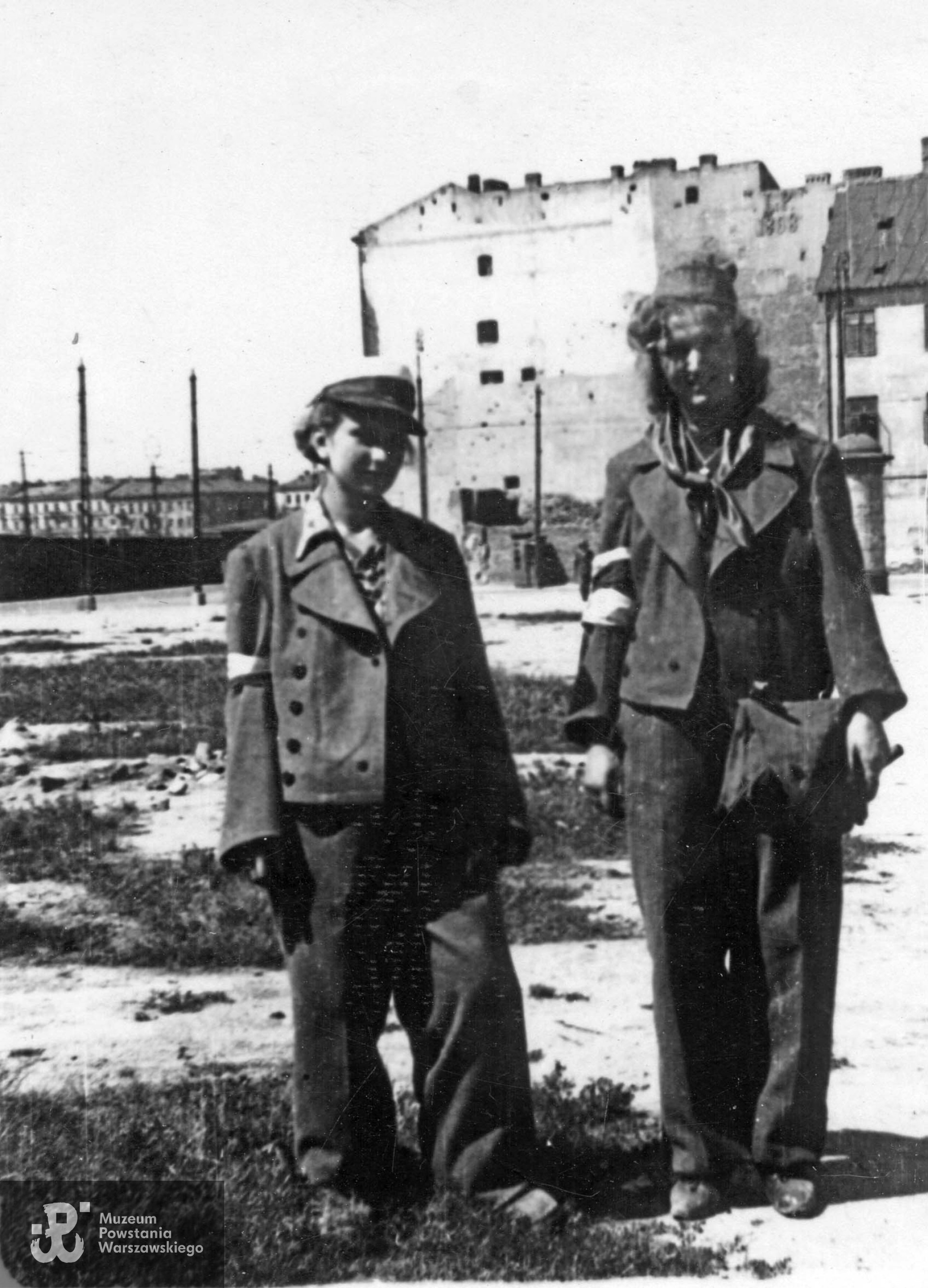 Fotografia autorstwa Zbigniewa Grochowskiego wykonana podczas Powstania Warszawskiego. Stare Miasto. Dwie łączniczki na skwerze przy ulicy Nowiniarskiej. W tle po prawej fragment budynku Franciszkańska 14 i ściana szczytowa kamienicy Bonifraterska 4. Daleko w tle (za murem byłego getta) od lewej domy: Muranowska 8, Muranowska 6, Muranowska 4, wylot ulicy Przebieg i fragment kamienicy Muranowska 2. Fot. ze zbiorów Muzeum Powstania Warszawskiego, sygn. MPW-IP/2695 (MPW-GI-4000/15/06).