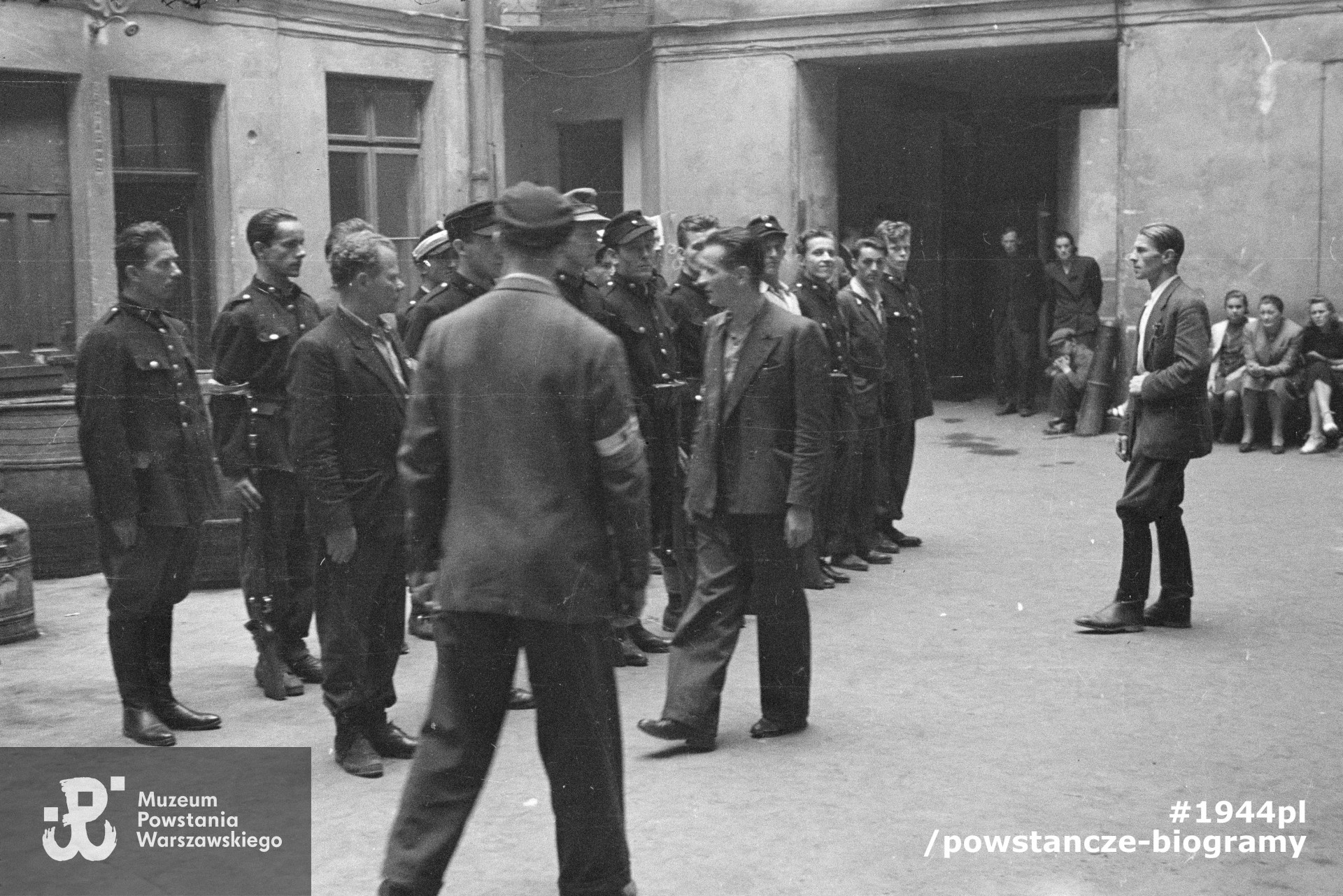 Fotografia z Powstania Warszawskiego. Śródmieście Północne. Zbiórka II plutonu kompanii "Koszta" na podwórzu kamienicy Jankowskiego przy ul. Moniuszki 12. Mjr Stefan Mich "Kmita" dokonuje przeglądu pododdziału. Na pierwszym planie (odwrócony tyłem) ppor. Stanisław Kuśpit "Kruk", pierwszy od prawej por. Roman Rozmiłowski "Zawada". Autorem zdjęcia jest Eugeniusz Lokajski. Fot. ze zbiorów Muzeum Powstania Warszawskiego, sygn. MPW-IN/160