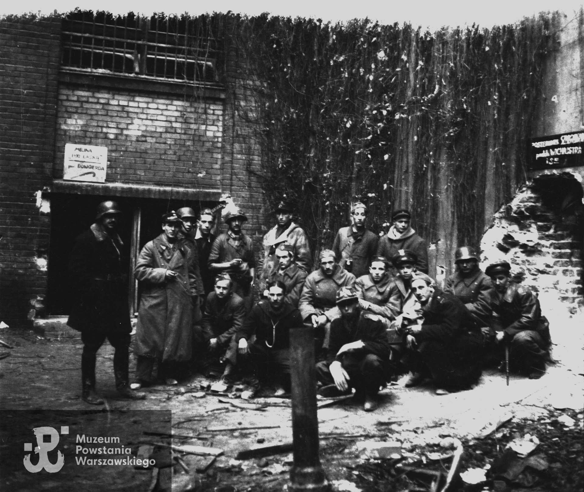 Zdjęcie grupowe 18 żołnierzy "Krybara", na murze po lewej stronie nad wejściem do budynku tabliczka: MELINA - "POD LASKĄ"- por. DOWGIRDA (por. Władysław Szella, 2. komp. Grupy Bojowej "Krybar"). Fot. ze zbiorów Muzeum Powstania Warszawskiego, sygn. MPW-IN/11129