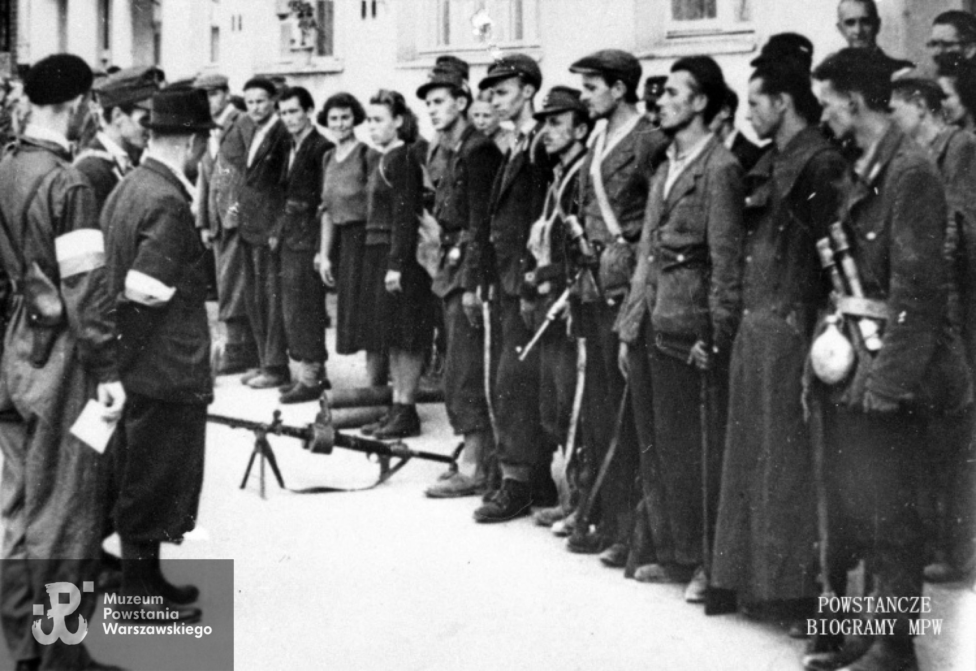 Zdjęcie filmowe z Powstania Warszawskiego. Mokotów, ulica Puławska 132. Dowódca pułku "Baszta" ppłk Stanisław Kamiński "Daniel" (w kapeluszu) dokonuje przeglądu oddziałów przybyłych z Lasów Chojnowskich. Pluton 3 kompanii K-1 pułku "Baszta". Od lewej tyłem: w berecie rtm. Lech Głuchowski "Jeżycki" dowódca dywizjonu "Jeleń", kpr. pchor. Tadeusz Krassowski "Start" zastępca dowódcy plutonu 3 kompanii K-1, ppłk Stanisław Kamiński "Daniel". W szeregu od prawej: czwarty w cyklistówce st. strz. Wacław Muszyński "Tyka", z chlebakiem st. strz. Janusz Różalski "Magnus", strz. Józef Kraszewski "Kot", kpr. Zygfryd Wiśniewski "Mściciel", łączniczka Danuta Kasprzycka-Deperazińska "Iwonka" (?) Tomasz Morawski stoi pierwszy z prawej.  Autor : Roman Banach "Świerk", zdjęcie filmowe. Prawa autorskie MPW, sygn. MPW-IH/1145