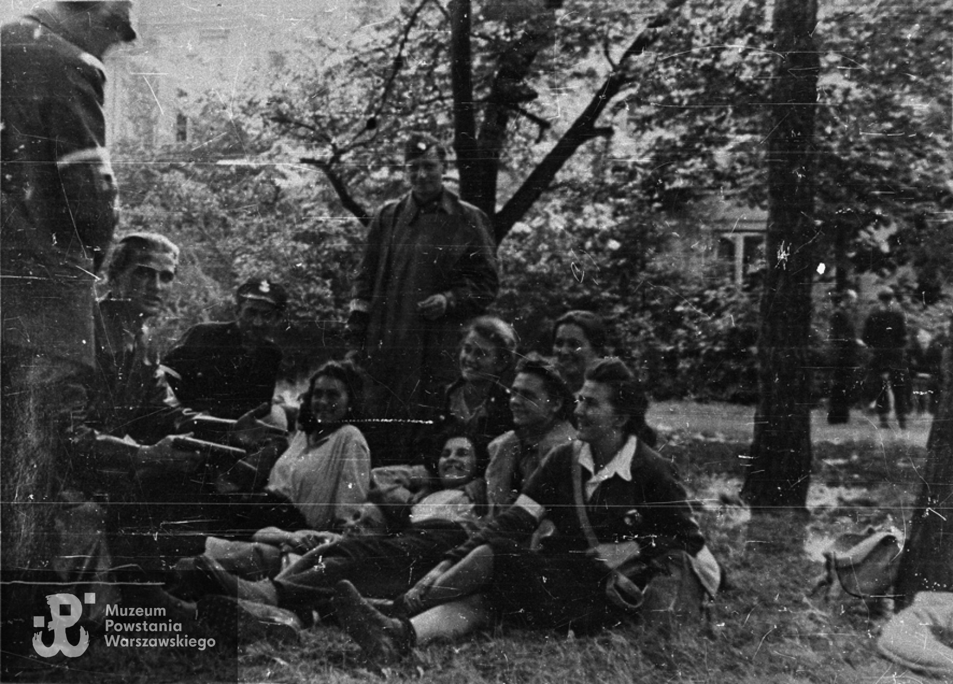 Fotografia z Powstania Warszawskiego. Stare Miasto, Ogród Krasińskich. Łączniczki, sanitariuszki i żołnierze batalionu "Chrobry I" w czasie wykładu o broni. Od lewej: z pistoletami strz. Bronisław Borowski "Słowik", kpr. "Gajda", Danuta Kamińska "Ewa"; stoi st. strz. "Leń"; leżą "Halszka", Krystyna Zapaśnik"Krystyna"; z rękami pod głową - N.N.; za nią - "Iga", "Jula" i "Iza". Fot. ze zbiorów Muzeum Powstania Warszawskiego, sygn. MPW-IN/8692. Autorem zdjęcia jest Zygmunt Sowiński "Ostoja"