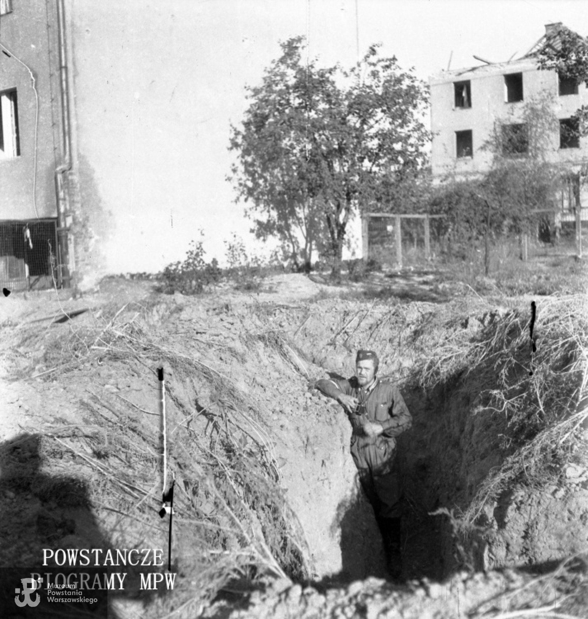 Fotografia z Powstania Warszawskiego. Mokotów. Rów dobiegowy przy budynku kwatery - Racławicka 17. W tle po prawej widoczny zniszczony budynek misji sióstr franciszkanek z Kaplicą Matki Boskiej Królowej Misji - Racławicka 14, róg Misyjna. Opis autora: "Dziura po nalocie na kwaterę naszą na Racławickiej". Fot. ze zbiorów Muzeum Powstania Warszawskiego, sygn. MPW-IN/6416