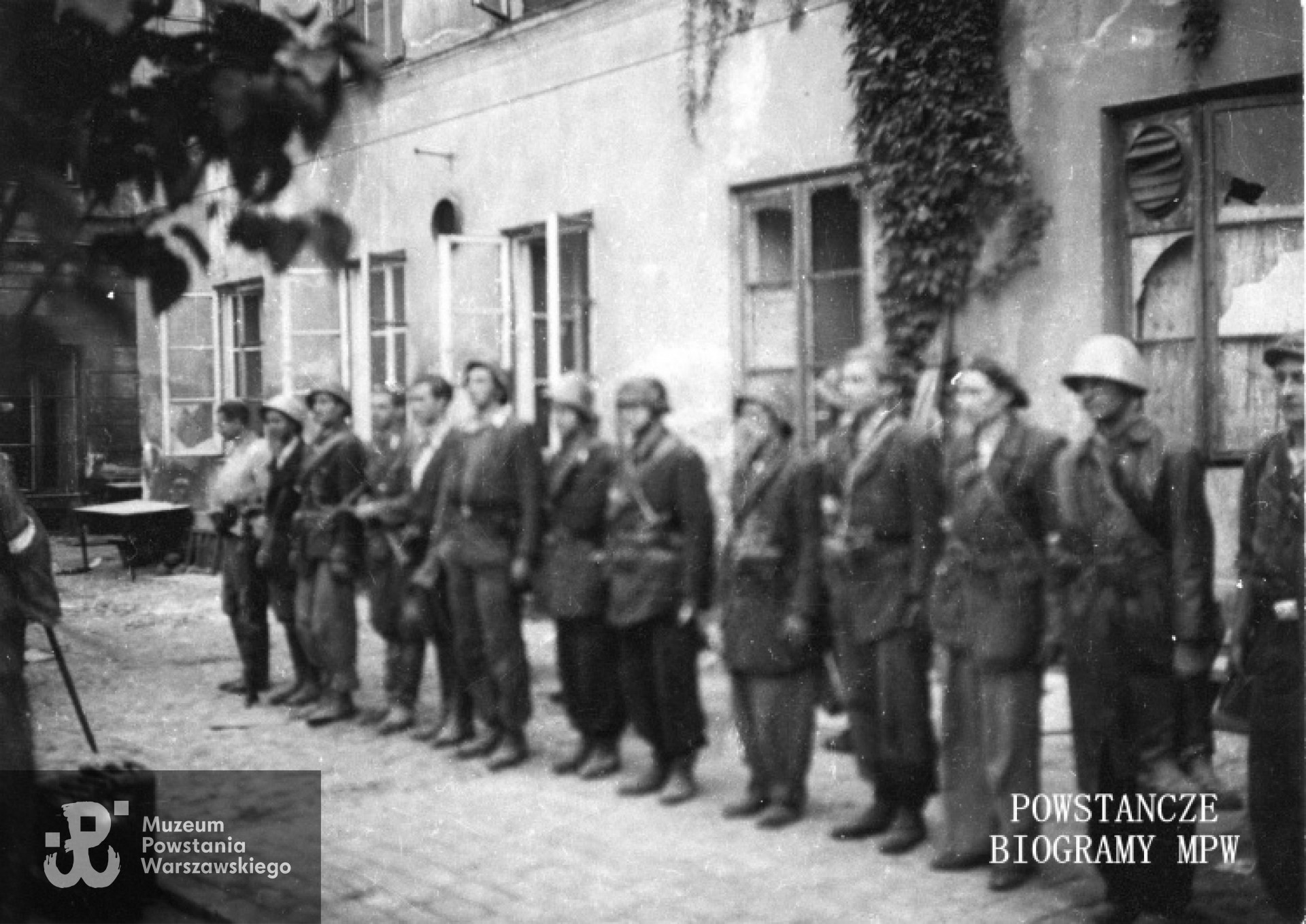 Fotografia z Powstania Warszawskiego. Zbiórka powstańców z plutonu "Rygla" zgrupowania "Bartkiewicz" po zakończonej akcji w podwórzu  zdobytej Komendy Policji. Po lewej widoczna częściowo postać kpt. Józefa Rybickiego "Andrzeja". W szeregu stoją od lewej: 1. ppor. NN "Grzegorz", 2. strz. Andrzej Bukraba "Żeglarz", 3.. sierż. pchor. Lucjan Kindlein "Lucjan", 4. kpr. pchor. Andrzej Gliński "Leszczyc", 5. NN., 6. strz. Mieczysław Śliwiński "Mietek", 7. kpr. pchor. Andrzej Strawiński "Chimera", 8. kpr. pchor. Józef Sowilski "Bohdan", 9. st. strz. Zbigniew Kurbiel "Krakus", 10. strz. Wiesław Nałęcz-Mroczkowski "Wiesiek", 11. NN, 12. kpr. pchor. Kazimierz Jesionowski "Jesion". Autor zdjęcia Irena Kummant-Skotnicka "Janina". MPW - sygn. MPW-IP/3828