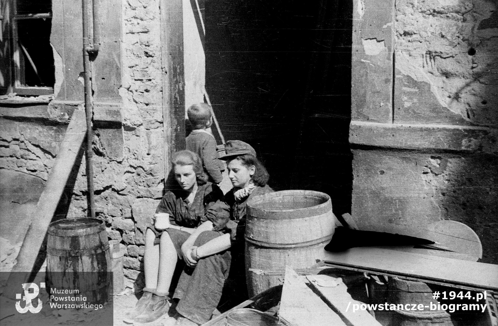 Fotografia z Powstania Warszawskiego. Sanitariuszki z kompanii "Anna" Batalionu "Gustaw": Danuta Magreczyńska "Danka" (po lewej) i Wanda Bagniewska "Wanda" na progu domu przy ulicy Ślepej. Za nimi chłopiec, jeden z mieszkańców kamienicy ukrywających się w piwnicach. Autorem zdjęcia jest Wiesław Chrzanowski "Wiesław". Fot. ze zbiorów Muzeum Powstania Warszawskiego, sygn. MPW-IN/3245