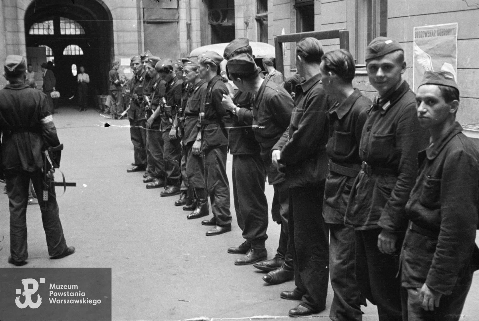 Fotografia z Powstania Warszawskiego. Śródmieście Północne. Apel III plutonu Oddziału Osłonowego Wojskowych Zakładów Wydawniczych (WZW) przy Oddziale VI Sztabu Komendy Głównej AK na podwórzu kamienicy Lewenfisza przy pl. Napoleona 1, róg ul. Boduena 2, róg ul. Sienkiewicza 1. Ujęcie w kierunku północnym. W tle widoczna brama wychodząca na ulicę Sienkiewicza. Pierwszy od prawej: st. strz. Tadeusz Wojda "Rzeźnik", strz. Włodzimierz Leo  "Bawół", kpr. pchor. Ryszard Wrzosek "Dick", (NN), kpr. pchor. Tadeusz Bruszewski "Sztubak". Na ścianie widoczny przedwojenny plakat PKO "Drogowskaz dobrobytu". Autorem zdjęcia jest Eugeniusz Lokajski "Brok". Fot. ze zbiorów Muzeum Powstania Warszawskiego, sygn. MPW-IN/714