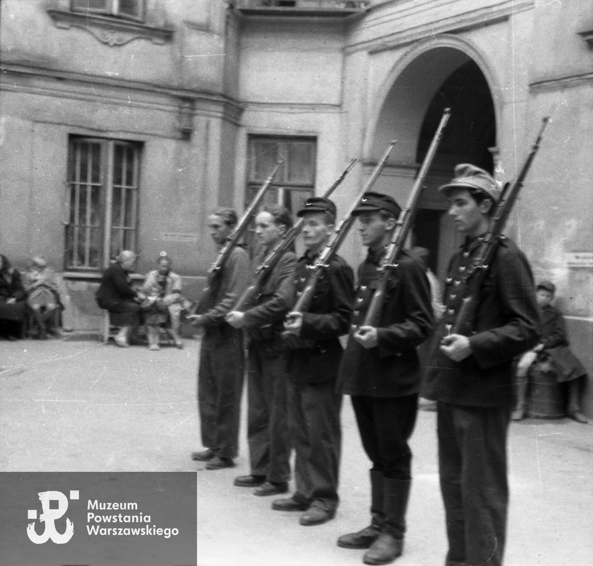 Fotografia z Powstania Warszawskiego. Śródmieście Północne. Grupa Powstańców z II plutonu kompanii "Koszta" podczas zbiórki na podwórzu kamienicy przy Moniuszki 12 róg Marszałkowska, za Powstańcami brama prowadząca na ulicę Moniuszki. Widoczni od prawej: strz. Paweł Kowalczyk "Andrzej", st. strz. Tadeusz Jankowski "Owa", NN, NN, st. strz. Zdzisław Kamiński "Czarny". Autorem zdjęcia jest Józef Namysłowski "Skiba". Fot. ze zbiorów Muzeum Powstania Warszawskiego, sygn. MPW-IN/6500