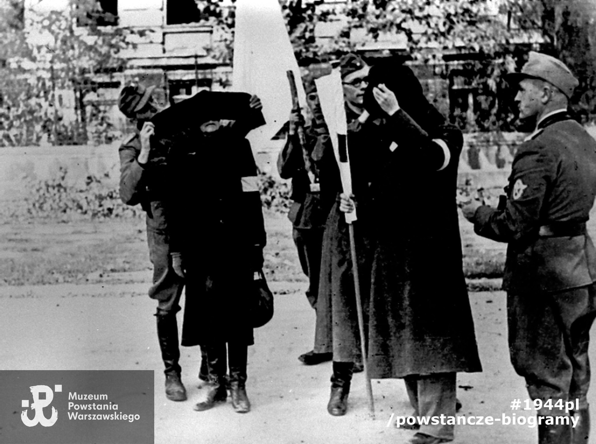 Fotografia z Powstania Warszawskiego. Śródmieście Południowe. Parlamentariusze polscy na ul. 6 Sierpnia (obecnie Nowowiejska) udają się na pertraktacje w sprawie kapitulacji Powstania. Moment zakładania członkom polskiej delegacji opasek na oczy. W tle gmach Politechniki Warszawskiej, fot. NN, Warszawa, 29 września 1944 r. Fot. ze zbiorów Muzeum Powstania Warszawskiego, sygn. MPW-IH/4270