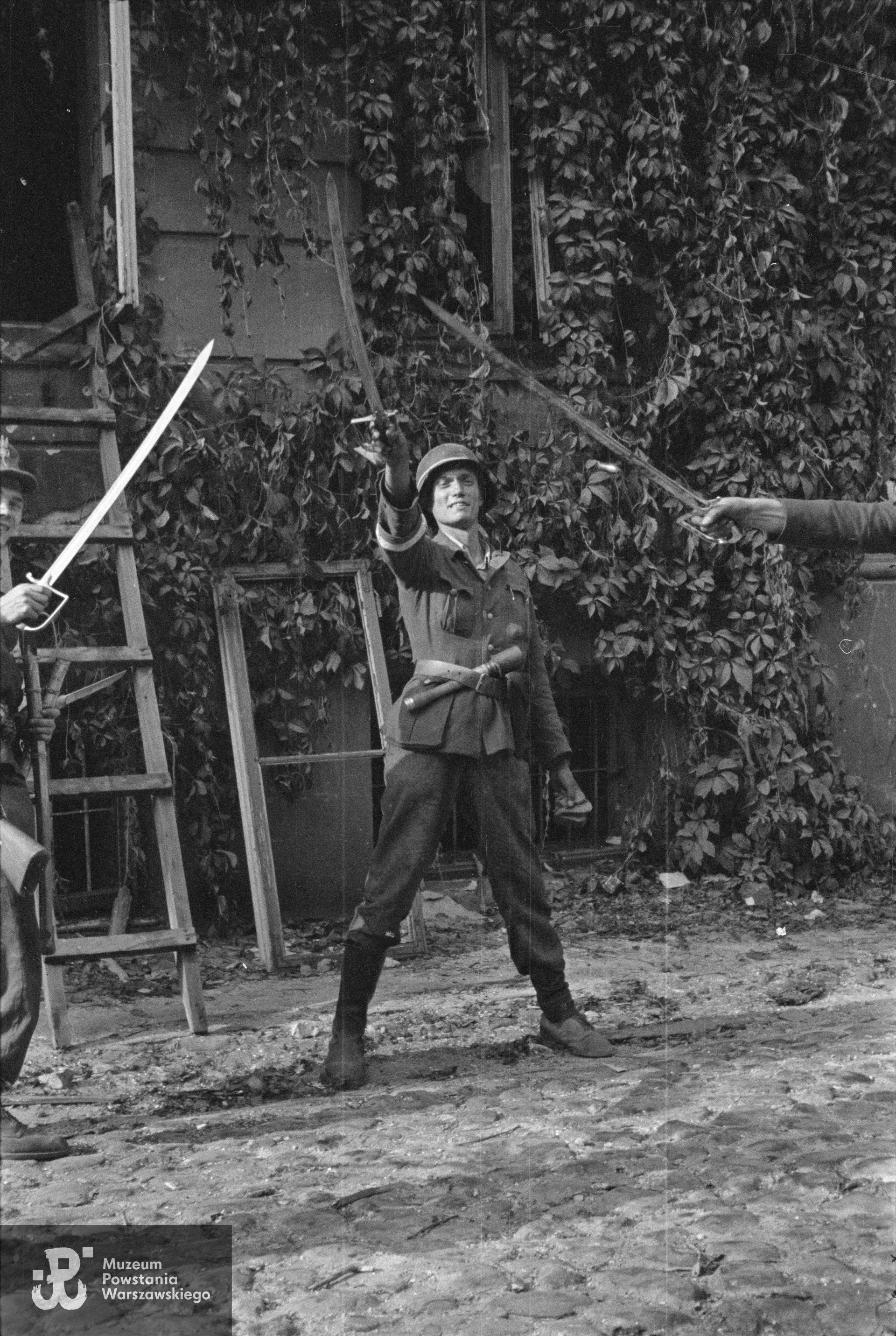 Fotografia z Powstania Warszawskiego: Śródmieście Północne - 23 sierpnia 1944 r. Powstańcy ze wzniesionymi polskimi szablami na dziedzińcu zdobytego gmachu Komendy Policji przy Krakowskim Przedmieściu 1; pośrodku stoi plut. pchor. Jerzy Tyczyński "Jur". Fot. autorstwa Eugeniusza Lokajskiego ps. "Brok". Ze zbiorów Muzeum Powstania Warszawskiego, sygn. MPW-IN/755.