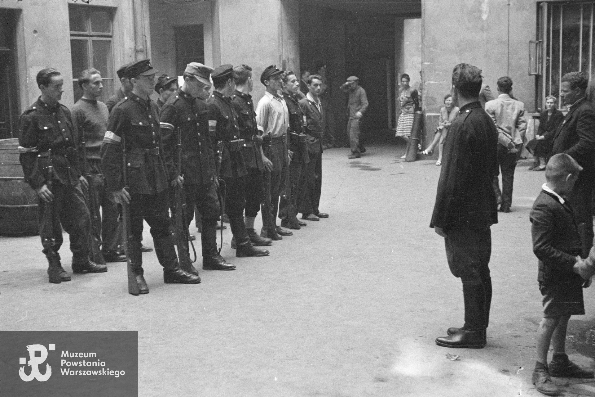  Fotografia z Powstania Warszawskiego. Śródmieście Północne. Zbiórka II plutonu kompanii "Koszta" na podwórzu kamienicy Jankowskiego przy ul. Moniuszki 12. W pierwszym szeregu od lewej: st. strz. Tadeusz Jankowski "Owa", strz. Paweł Kowalczyk "Andrzej", strz. Jerzy Baranowski "Wróbel" (w białej koszuli); w drugim szeregu od lewej: strz. Stanisław Zieliński "Zielony", st. strz.  Zdzisław Kamiński "Czarny".  Po prawej przed frontem plutonu stoi prowadzący musztrę plut. Edmund Klimaszewski "Jerzy". Fot. ze zbiorów Muzeum Powstania Warszawskiego, sygn. MPW-IN/159
