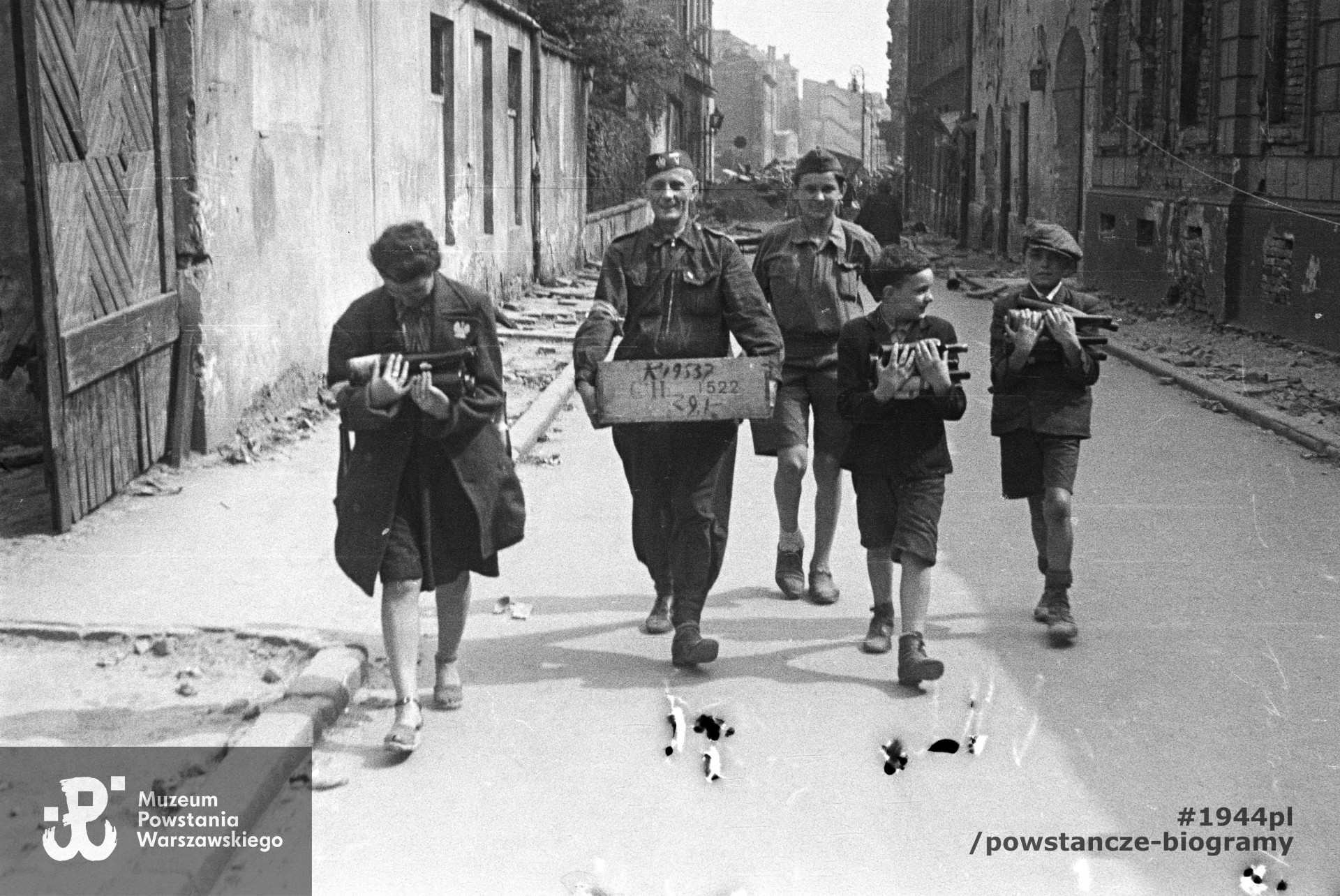 Fotografia z Powstania Warszawskiego. Ul. Warecka, widok w kierunku Nowego Światu. Na pierwszym planie grupa powstańców idących środkiem ulicy.  Po prawej budynki Warecka 3 i Warecka 1 (róg Nowy Świat 51), po lewej zabudowania na tyłach kamienicy Mikulskiego przy Nowym Świecie 53. W tle ruiny kamienic przy ul. Ordynackiej. Według identyfikacji dokonanej przez p. Marka Szandorowskiego chłopiec idący z prawej strony to on. Autorem zdjęcia jest Tadeusz Bukowski "Bończa". Fot. ze zbiorów Muzeum Powstania Warszawskiego, sygn. MPW-IN/11989