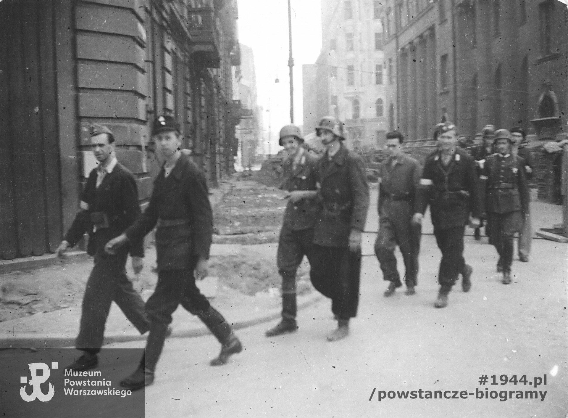 Fotografia z Powstania Warszawskiego. Idący dwójkami powstańcy z plutonu "Rygla" zgrupowania "Bartkiewicz", skręcający z ul. Świętokrzyskiej w Szkolną, ujęcie w kierunku wsch. Idą od lewej: kpr. z cenz. Jerzym Jenike "Zwierz", kpr. pchor. Janusz Sawicki "Zabawa", kpr. pchor. Andrzej Gliński "Leszczyc", strz. Władysław Mydell "Waldemar" st. strz. Zbigniew Kurbiel "Krakus", kpr. pchor. Andrzej Strawiński "Chimera". Na wprost wylot ulicy Świętokrzyskiej. Po lewej na pierwszym planie kamienica Świętokrzyska 36/38/40 róg Jasna 11 róg Szkolna 2. Po prawej gmach PKO Świętokrzyska 31/33 róg Jasna 9, dalej widoczny narożnik kamienicy Świętokrzyska 27 róg Jasna 16. Ze zbiorów Muzeum Powstania Warszawskiego, sygn.  MPW-IP/3845
