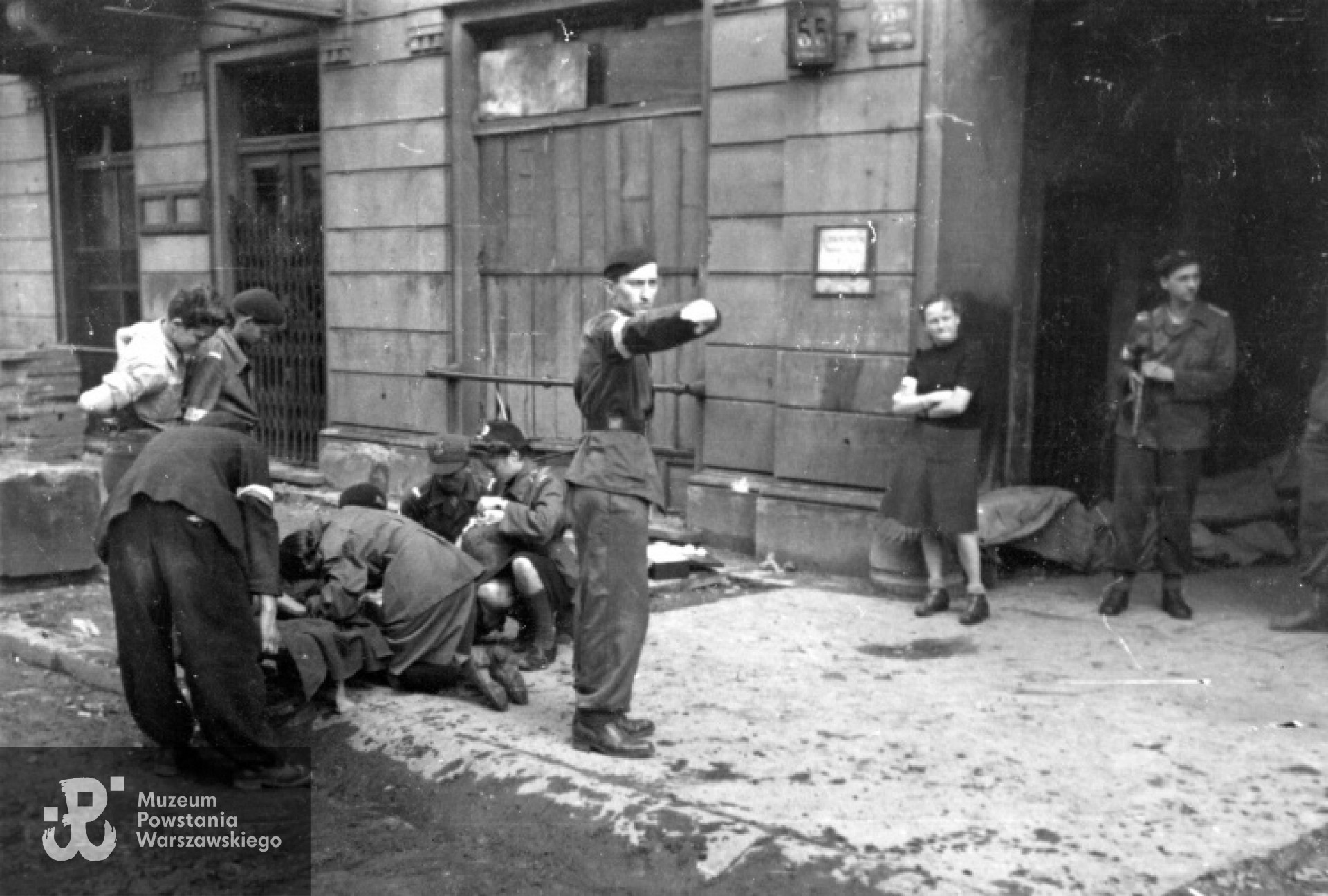 10 sierpnia 1944 : patrol sanitarny opatruje rannego powstańca z 2 Harcerskiej Baterii Artylerii Przeciwlotniczej "Żbik" przy ul. Chmielnej 55. Na noszach leży postrzelony kan. Ryszard Głuchowski "Jaworzak" Stoją od lewej: 1. pochylony sierż. Henryk Łyżwiński "Czarny" 2. kpr. pchor. Andrzej Rażniewski "Krzycki" 3. kpr. pchor. Piotr Osiński "Kaczanowski" 4. kpr. Tadeusz Błażyński "Świda" Klęczą od lewej : 1. kpr. pchor. Adam Bóbr „Jaxa” 2. kpr. pchor. Jerzy Bogdanowicz "Hercuń" 2. kpr. sanit. Elżbieta Tatarkiewicz "Żabska" 3. strz. sanit. Maria Jezierska "Bogdańska" Stoją w bramie: strz. łączn. Jadwiga Mundkowska "Dina Pękałówna" kan. Juliusz Smardzewski "Cezar". Autorem zdjęcia jest Joachim Joachimczyk. Zbiory fototeki MPW, prawa autorskie MPW. Sygn. <a href=" http://www.1944.pl/galerie/fototeka/foto/1812/kategoria/76"> MPW-IP/2666</a>