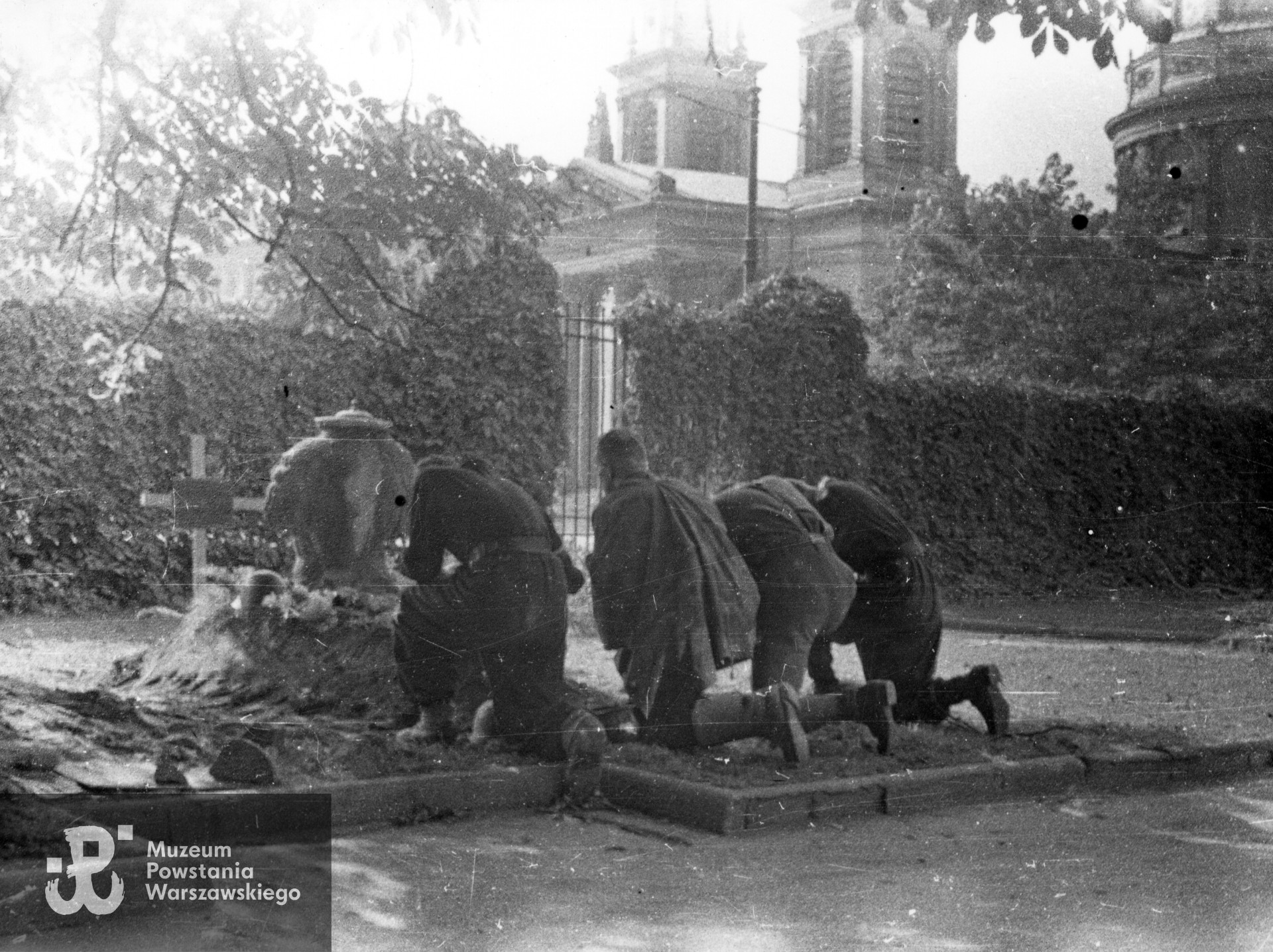 Fotografia z Powstania Warszawskiego. Śródmieście Południowe, pl. Trzech Krzyży. Grób strz. Witolda Hermanowskiego "Świta" z 1. plutonu "Kłosa" 3. kompanii "Redy" batalionu "Miłosz" na terenie Instytutu Głuchoniemych i Ociemniałych przy pl. Trzech Krzyży 4/6. Klęczą od lewej: pchor. Juliusz Pietrzykowski "Teddy", kpr. pchr. Stanisław Szabłowski "Witeź", "Grom", strz. Jerzy Ryszard Rosiak "Pantera". Ujęcie w kierunku północno - zachodnim. W tle widoczna bryła kościoła św. Aleksandra. Autorem zdjęcia jest Wojciech Marczyński "Bogusław". Fot. ze zbiorów Muzeum Powstania Warszawskiego, sygn. MPW-IK/2327