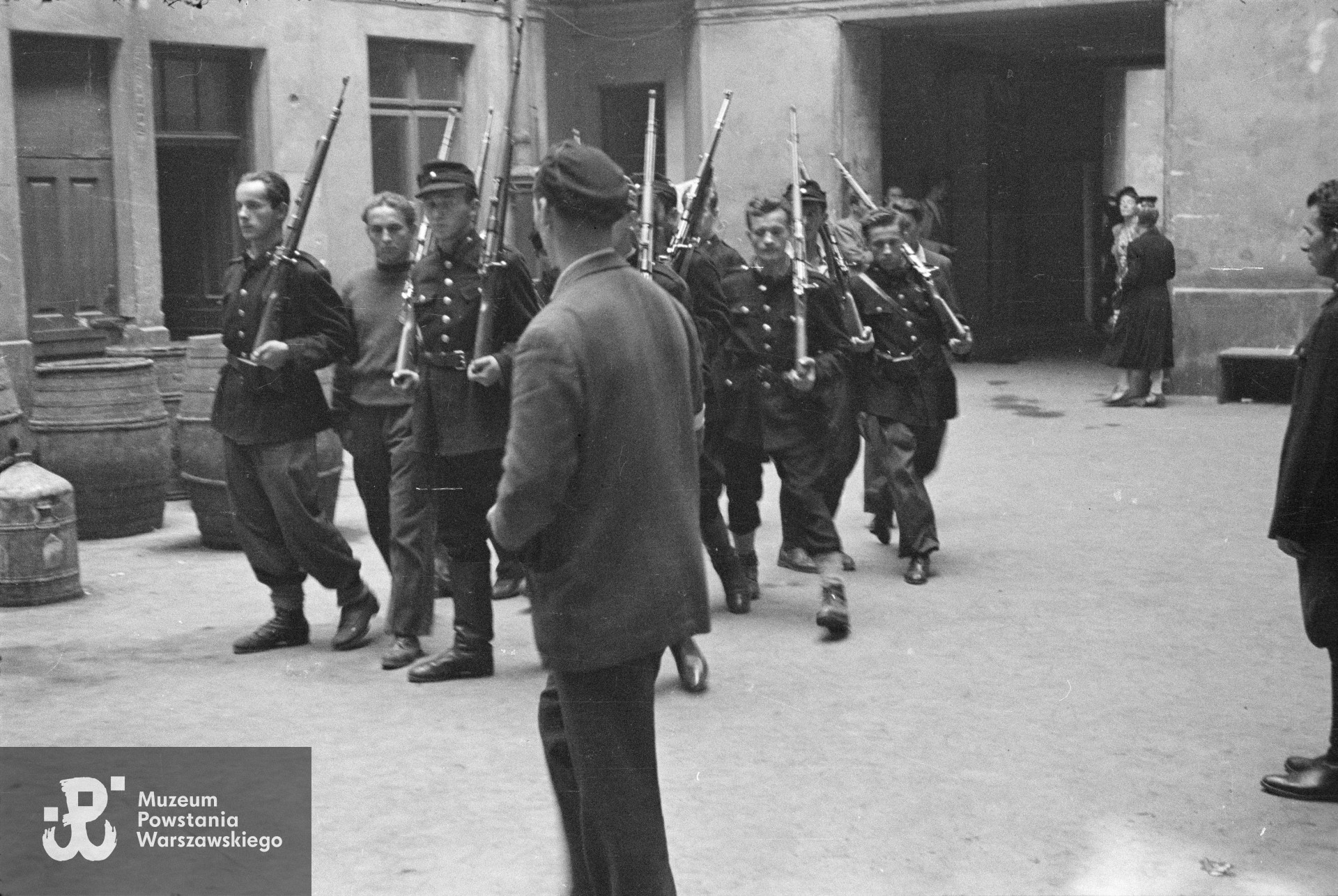  Musztra II plutonu kompanii "Koszta" na podwórzu kamienicy Jankowskiego przy ul. Moniuszki 12. Na pierwszym planie (odwrócony tyłem) stoi ppor. Stanisław Kuśpit "Kruk", nieopodal niego stoi prowadzący musztrę plut. Edmund Klimaszewski "Jerzy"; widoczna na fotografii pierwsza trójka od prawej: st. strz. Tadeusz Jankowski "Owa", st. strz. Zdzisław Kamiński "Czarny", strz. Stanisław Zieliński "Zielony". Fot. ze zbiorów Muzeum Powstania Warszawskiego, sygn. MPW-IN/158