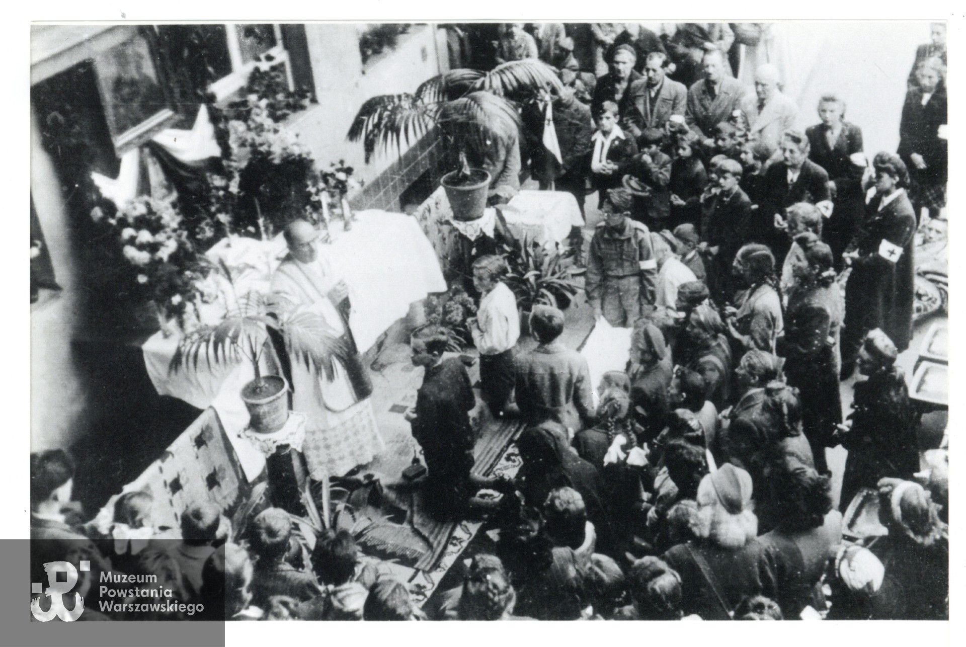 Fotografia z Powstania Warszawskiego. Sierpień 1944 r.,  msza św. polowa odprawiana na podwórku bloku przy ul. Mickiewicza 25. Łącznik Andrzej Chmielewski "Winnetu"  widoczny przy ołtarzu: ubrany w harcerski mundur, z powstańczą biało-czerwoną opaską na lewym ramieniu, klęczy trzymając obrus przy którym nastąpi udzielanie komunii wiernym.Fot. ze zbiorów Muzeum Powstania Warszawskiego, dar p. Zbigniewa Chmielewskiego.