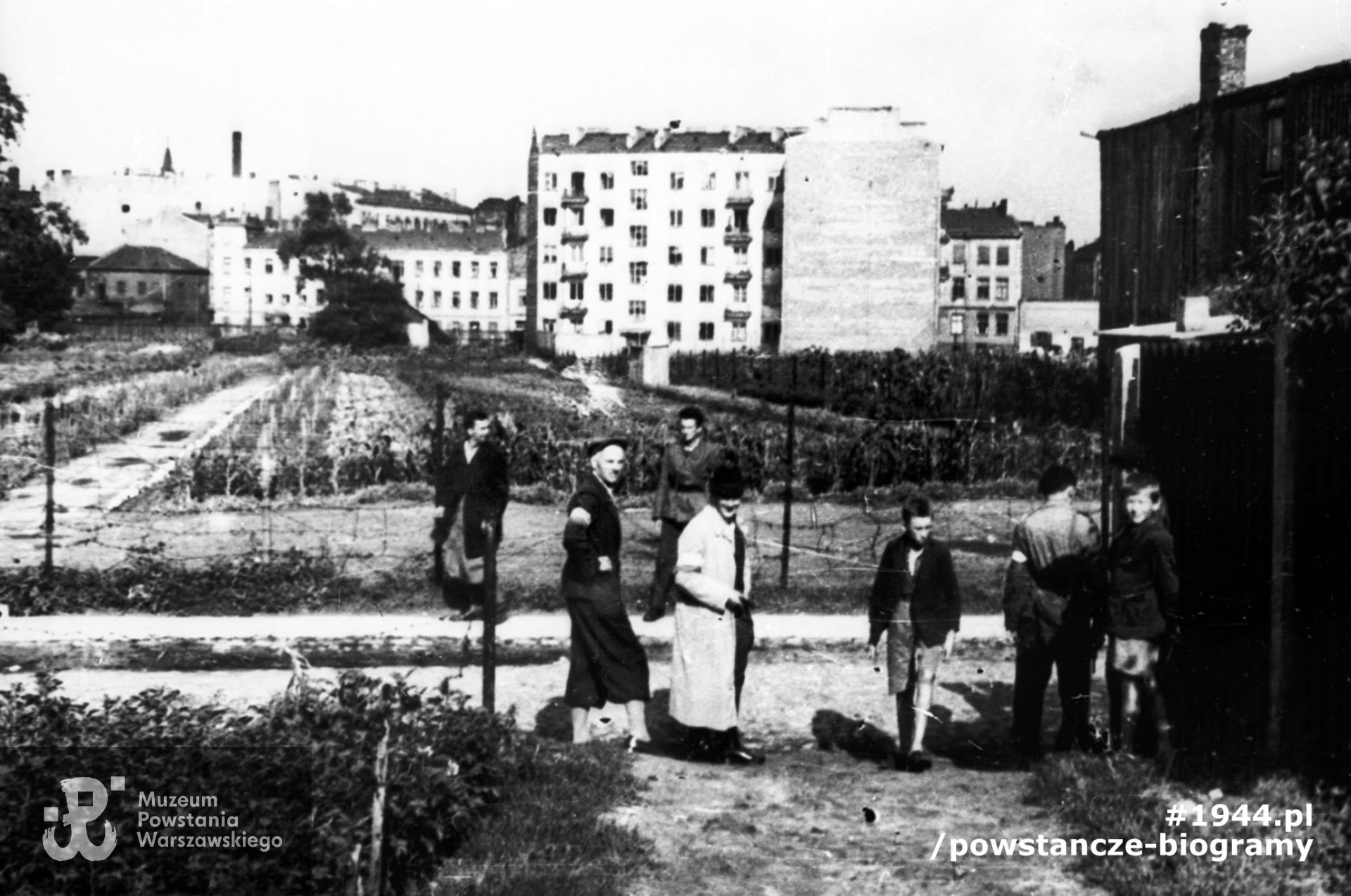  Fotografia z Powstania Warszawskiego. Wola. Godzina "W". Grupa powstańców z Batalionu "Pięść" stojąca przy wylocie ul. Długosza na Karolkową, oczekująca na koncentrację w rejonie cmentarza ewangelicko-augsburskiego. Po lewej, ubrany w spodnie pumpy, stoi mjr Alfons Kotowski "Okoń", dowódca Batalionu, w jasnym płaszczu ppor. Stanisław Bąk "Burza". Obok nich stoją chłopcy. Ujęcie w kierunku wschodnim. W tle widoczne budynki przy zbiegu ul. Okopowej i ul. Wolność; po lewej kamienica przy Okopowej 30 róg Wolność 21, w środku czteropiętrowa kamienica przy Okopowej 33. Autorem zdjęcia jest Stefan Bałuk. Ze zbiorów Muzeum Powstania Warszawskiego, sygn. MPW-IH/3408. Prawa autorskie MPW