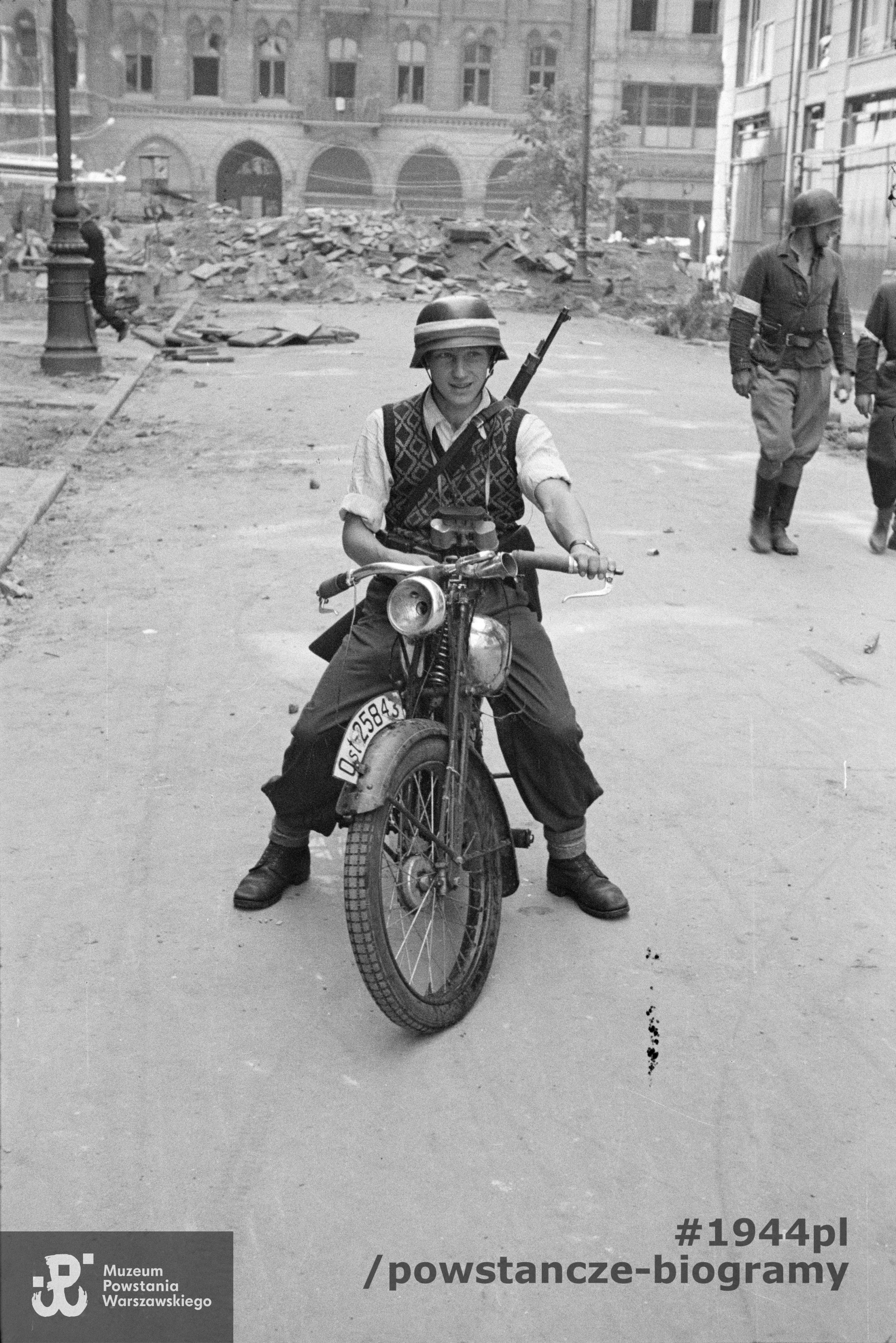 Fotografia z Powstania Warszawskiego. Śródmieście Północne. Kpr. pchor. Antoni Tuleja "Niedźwiedź" z kompanii "Koszta" siedzący na motocyklu na ul. Moniuszki. Ujęcie w kierunku zachodnim. Na drugim planie po prawej dwóch rozmawiających powstańców i narożnik kamienicy Jankowskiego przy ul. Moniuszki 12. W tle widoczna barykada zamykająca wylot na ulicę Marszałkowską, za nią kamienice braci Zamboni przy ul. Marszałkowskiej 127 (po lewej) oraz fragment sąsiedniej pod nr 129. Autorem zdjęcia jest  Eugeniusz Lokajski "Brok". Ze zbiorów Muzeum Powstania Warszawskiego,  sygn. MPW-IN/148