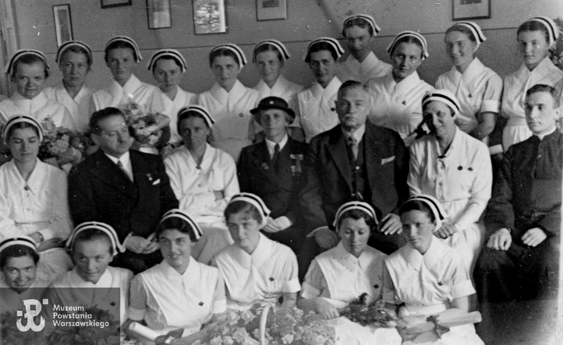 Fotografia z okresu międzywojennego. Zdjęcie pamiątkowe po złożeniu przyrzeczenia przez absolwentki Szkoły Pielęgniarstwa PCK przy ul. Smolnej 6.  W najwyższym rzędzie: trzecia od lewej - Halina Glińska (późniejsza Bukowska), w środkowym rzędzie: drugi od lewej naczelnik Krakowski z Ministerstwa Opieki Społecznej, od prawej: kapelan Szkoły Pielęgniarstwa PCK ks. Godziński, druga od prawej dyrektorka szkoły Małgorzata Żmudzka, obok niej dyrektor szpitala PCK prof. Bolesław Szarecki. Autorem zdjęcia jest Tadeusz Bukowski. Fot. ze zbiorów Muzeum Powstania Warszawskiego, sygn. MPW-IP/10421 (P/8995/18)