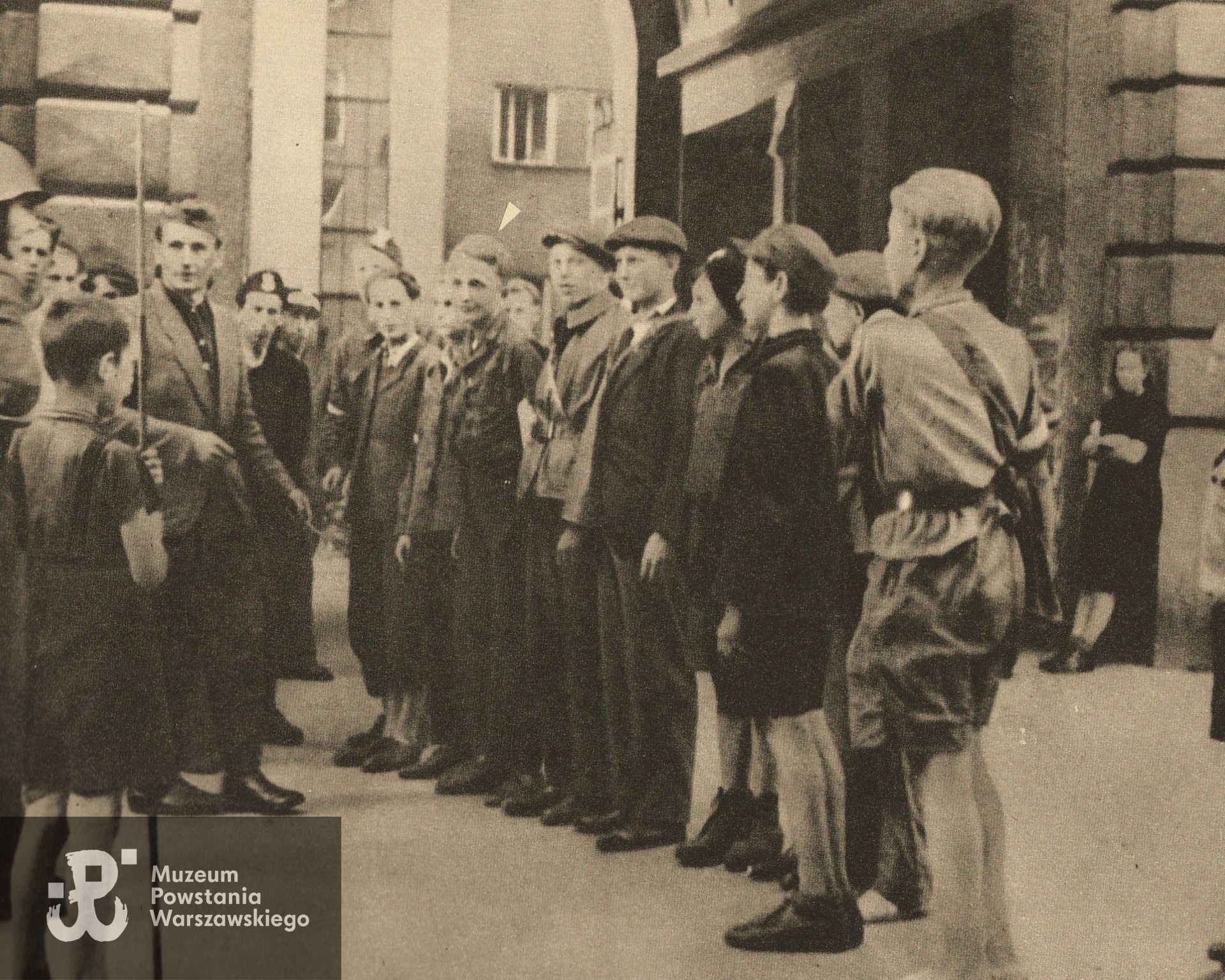 Fotografia z Powstania Warszawskiego. Powiśle. Zbiórka powstańczego oddziału przed bramą domu przy ul. Smulikowskiego 1/3. Jerzy Gaździcki "Gołąb" oznaczony strzałką. Opis do zdjęcia: "Składanie przysięgi AK na Smulikowskiego przy dowództwie III Zgrupowania "Konrad". Ze zbiorów p. Jerzego Gaździckiego ps. Gołąb